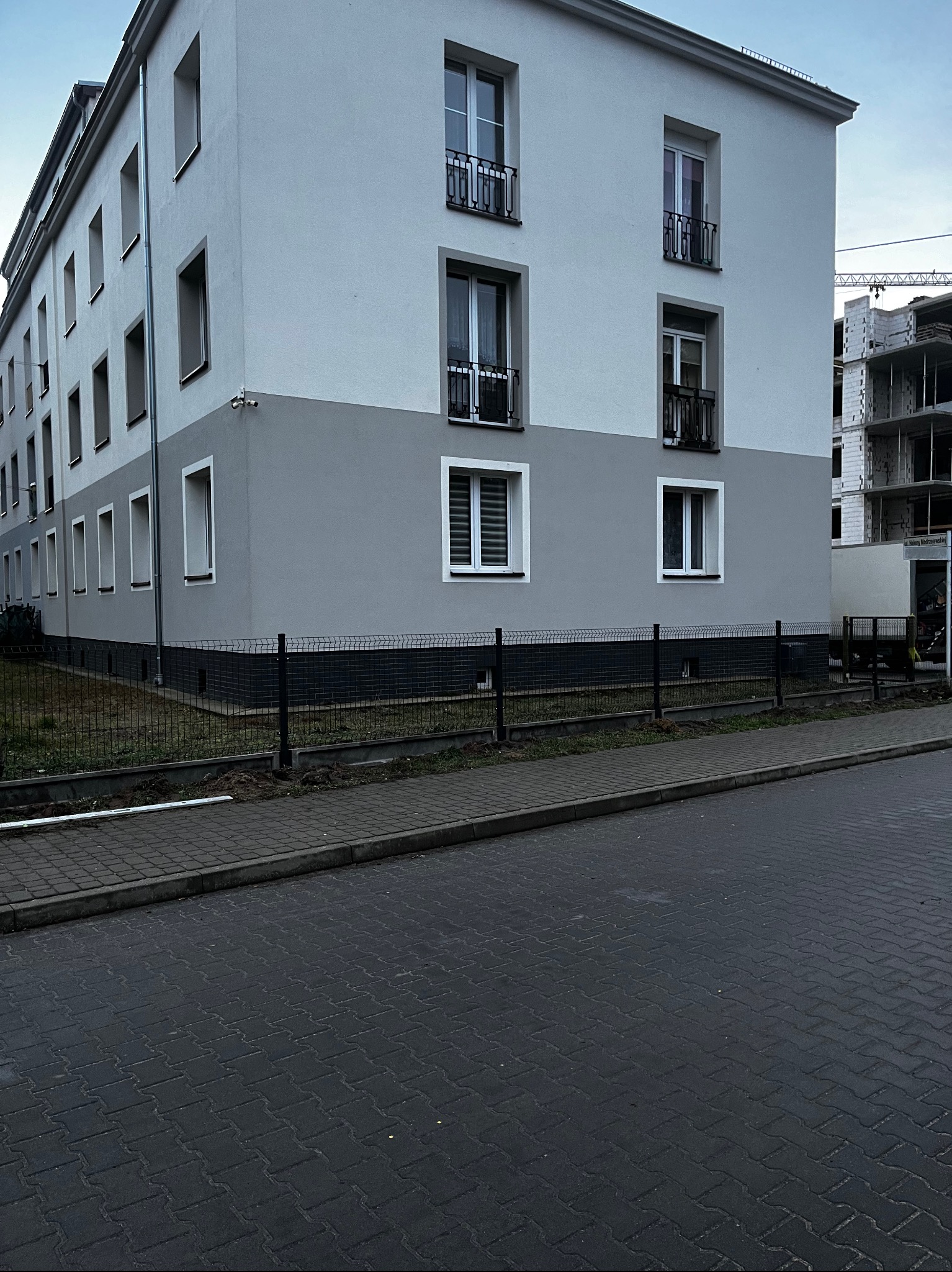 Dwukondygnacyjny budynek z szarą dolną częścią elewacji i czarnym ogrodzeniem panelowym. W tle widoczna konstrukcja w trakcie budowy.