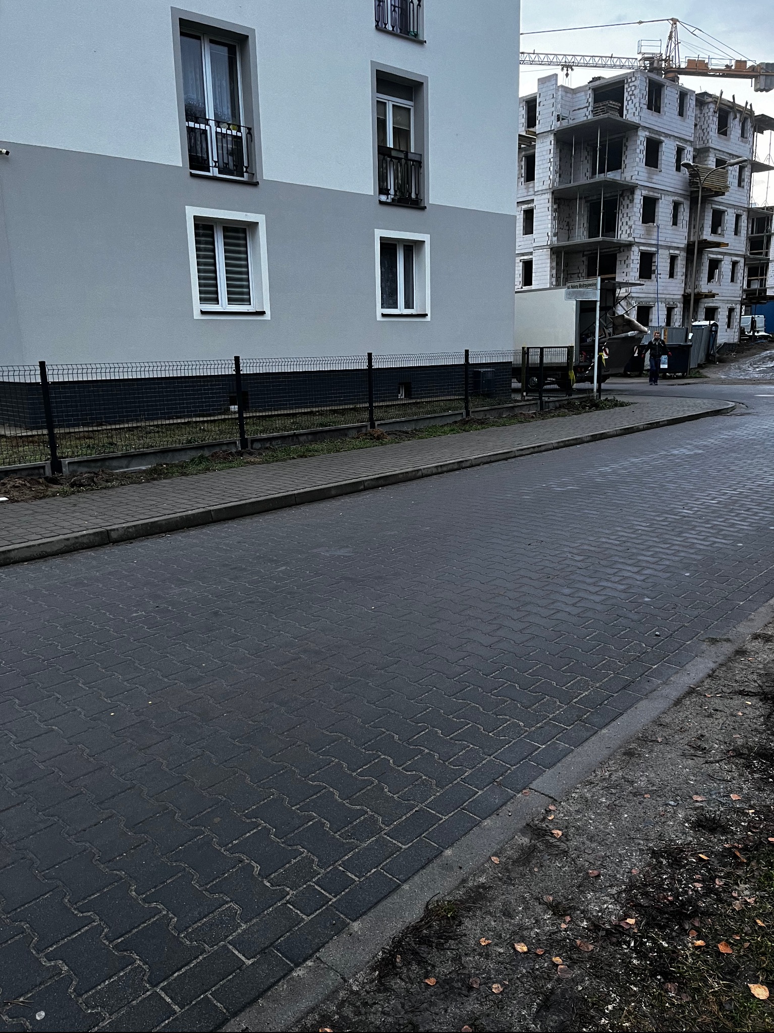 Nowoczesny budynek z szarą elewacją i oknami PCV, ogrodzony niskim, czarnym płotem. W tle budowa z dźwigiem. Ujęcie z poziomu chodnika z kostki.