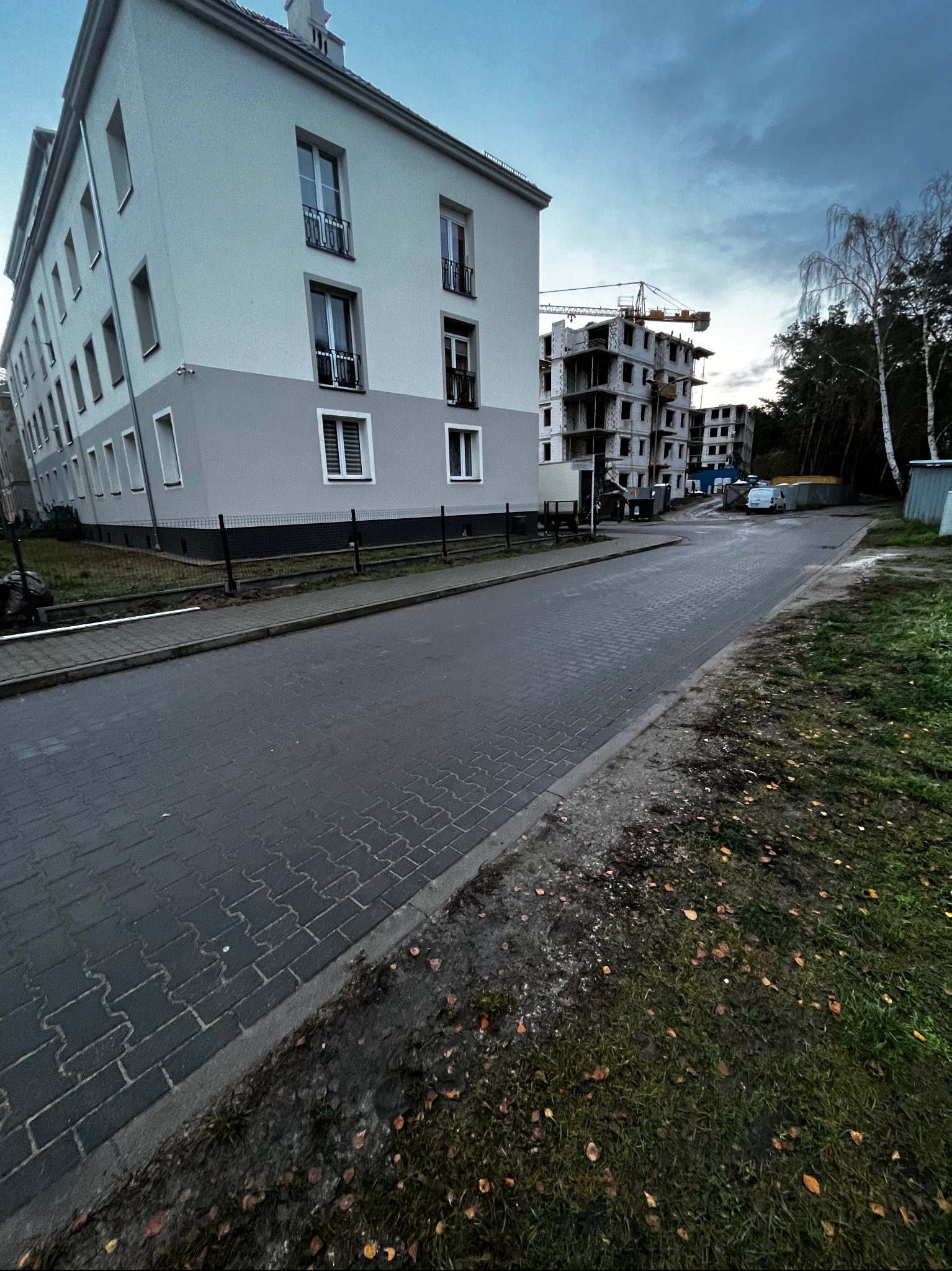 Nowoczesny budynek wielorodzinny z szarą elewacją i czarnymi balkonami, w tle budowa z dźwigiem, widok z poziomu ulicy w pochmurny dzień.