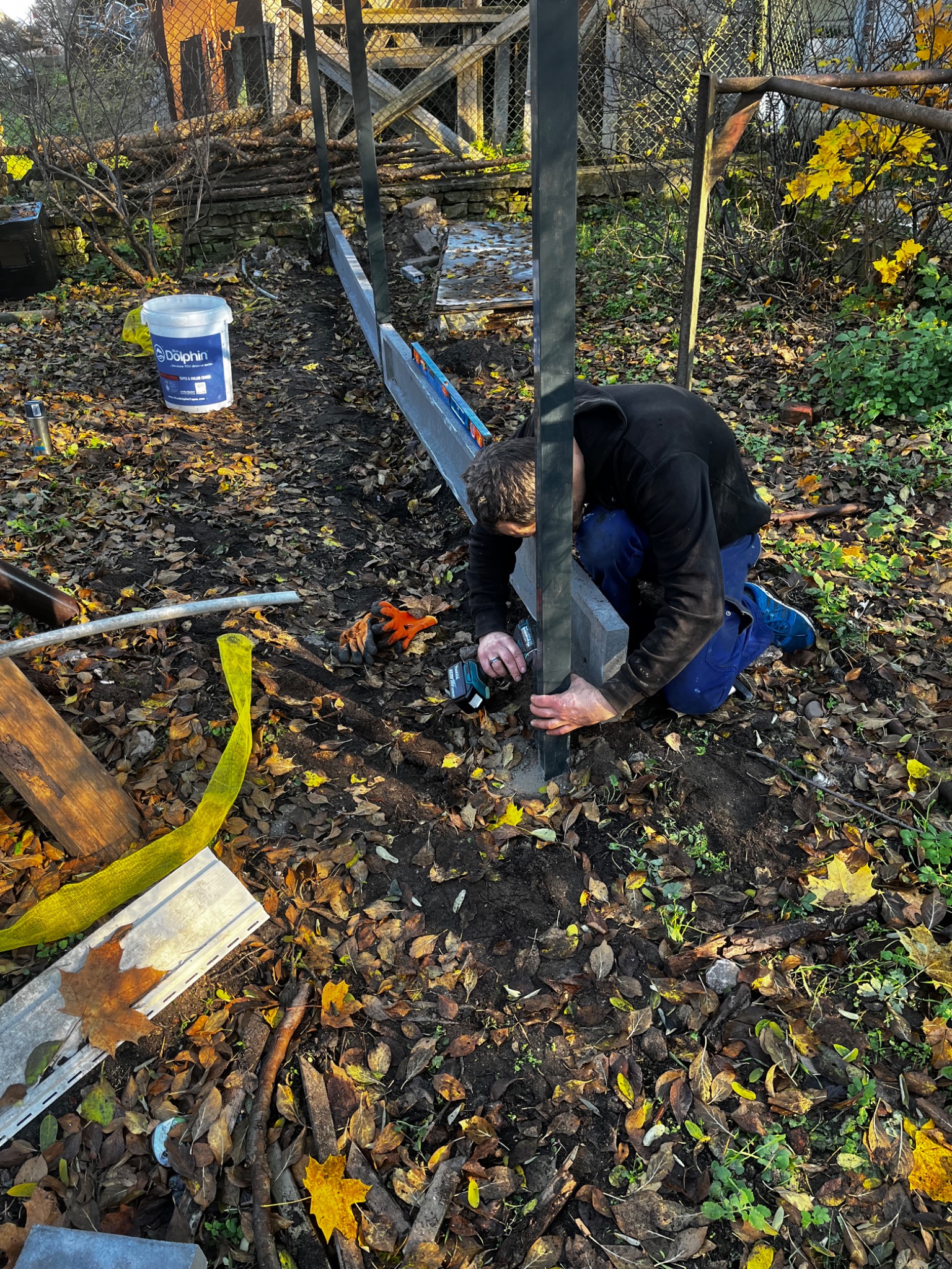 Pracownik w trakcie montażu słupków ogrodzeniowych na zewnątrz, używając wkrętarki. Widoczne narzędzia, poziomica i wiadro z farbą. Jesienne liście na ziemi.