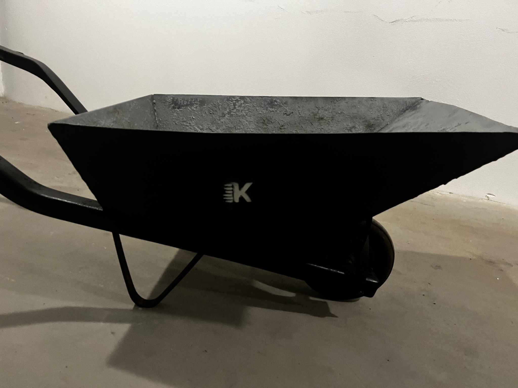 Czarna taczka z logo EK, ustawiona na betonowej posadzce, na tle białej ściany. Widoczny fragment uchwytu i koła. Minimalistyczne ujęcie.