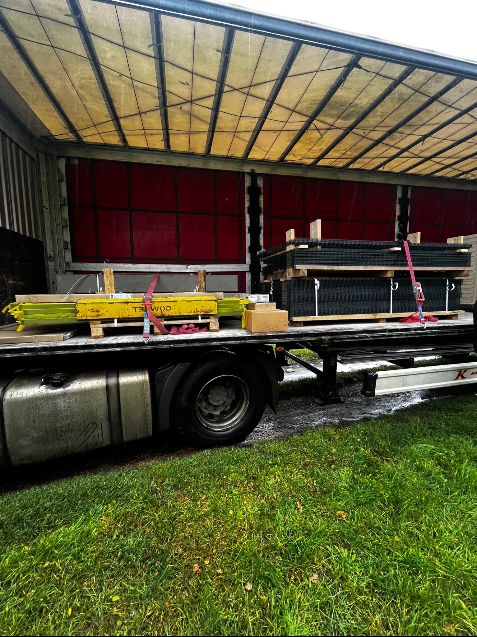 Transport ogrodzenia panelowego na paletach w naczepie ciężarówki, zabezpieczone pasami. Widoczne elementy żółte i czarne. Trawa w tle.