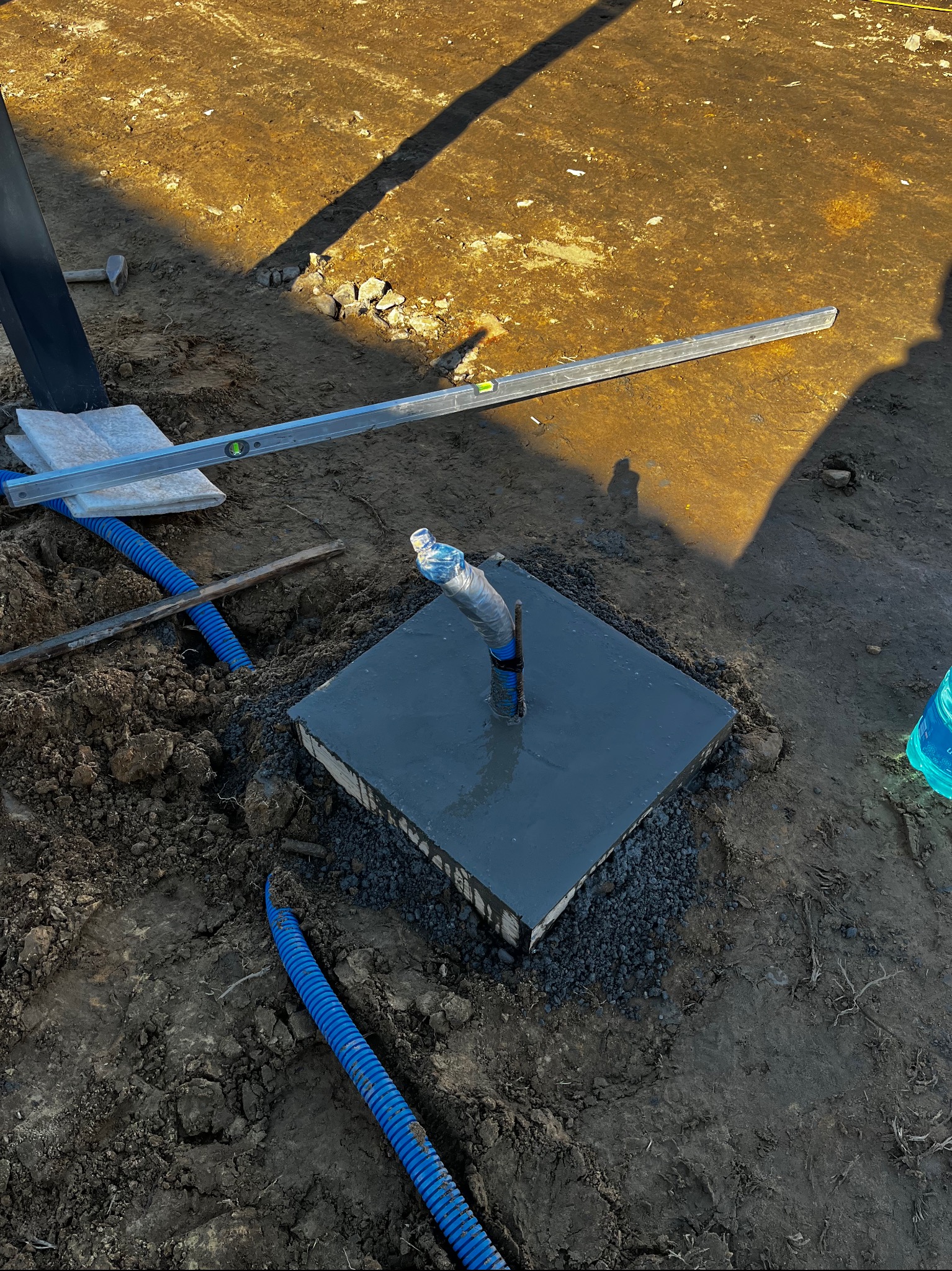 Świeżo wylany, kwadratowy fundament pod słupek ogrodzeniowy z wystającą rurą, poziomica i niebieskie rury karbowane w ziemi. Widoczne narzędzia i cień słupka.