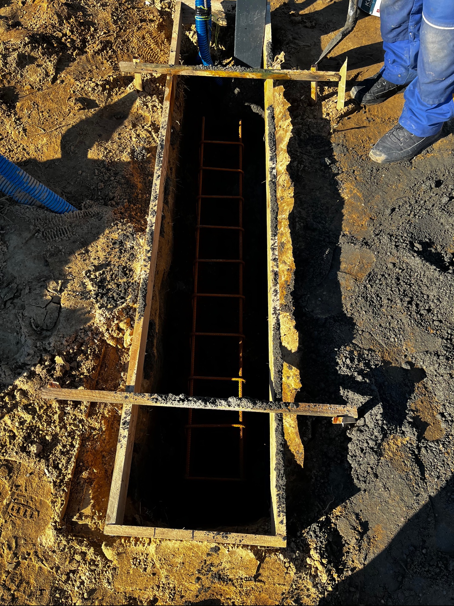 Wykop z deskowaniem i zbrojeniem, przygotowany pod fundament. Widoczne fragmenty węży i narzędzi. Praca w toku, ziemia i piasek wokół wykopu.