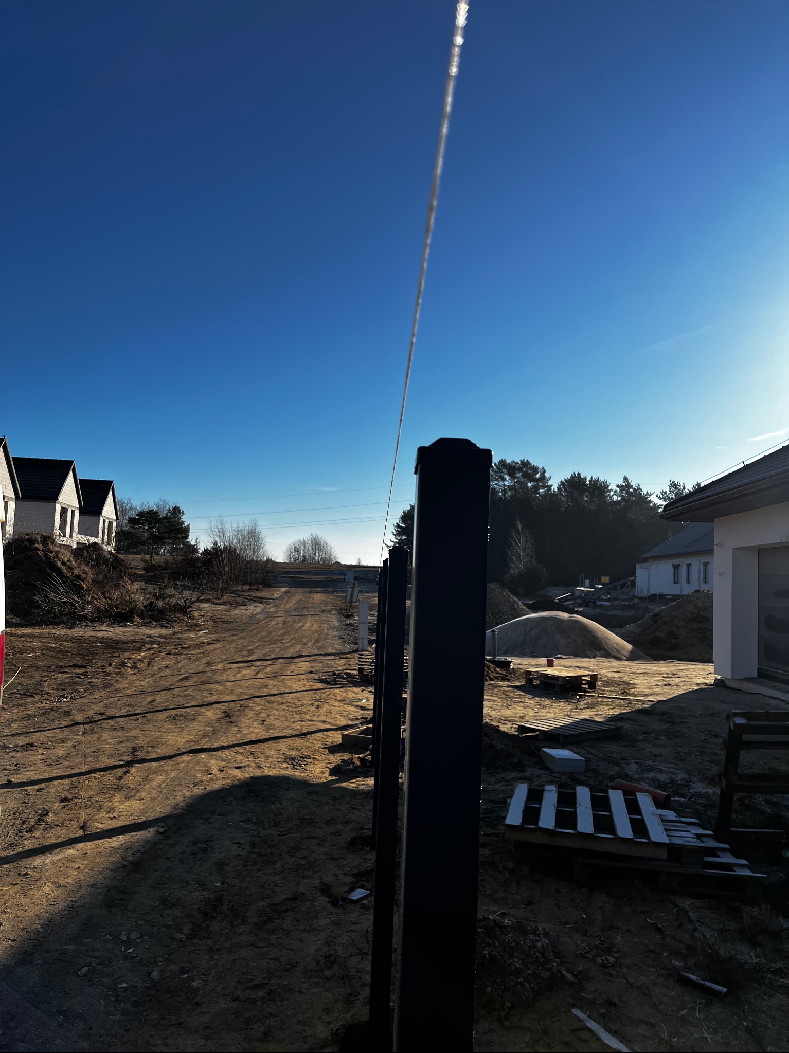 Czarne słupki ogrodzeniowe w trakcie montażu na tle placu budowy z domami i błękitnym niebem. Widoczne palety i piasek.