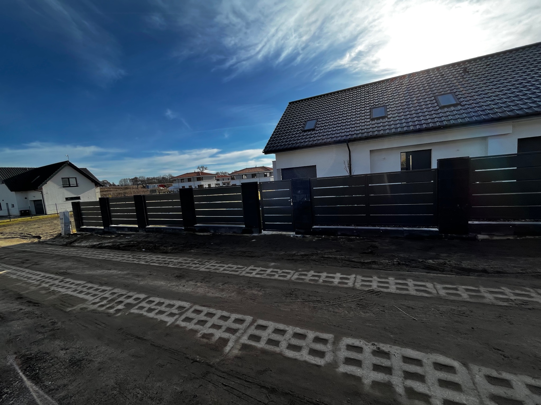 Nowoczesne ogrodzenie panelowe w kolorze antracytowym, z poziomymi przęsłami, na tle domu z ciemną dachówką i błękitnego nieba z chmurami. Widoczne ślady opon na ziemi.