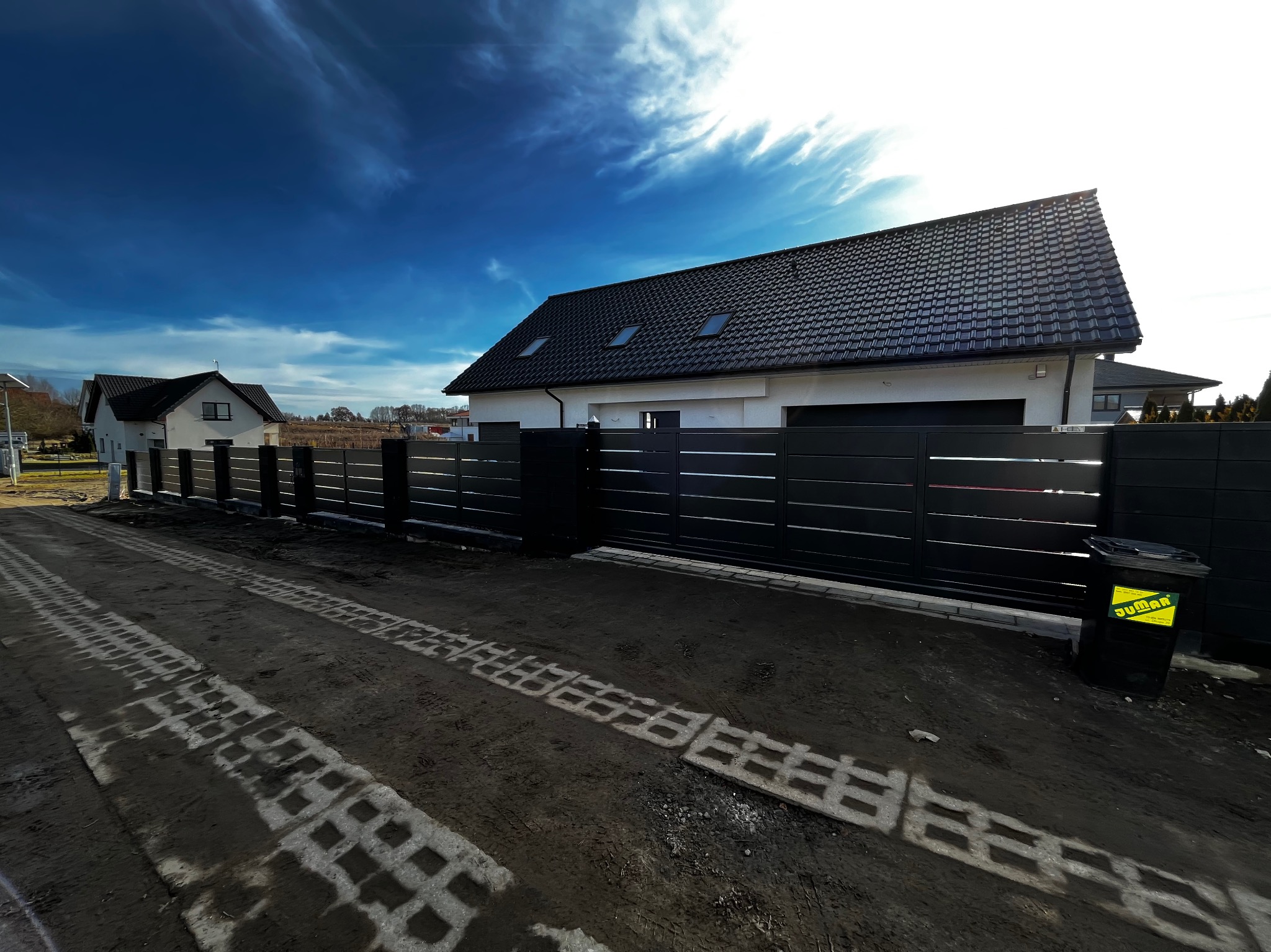 Nowoczesna, pozioma, czarna brama wjazdowa z przesuwnym skrzydłem, widoczna z zewnątrz posesji. Na pierwszym planie ślady po kostce ażurowej.