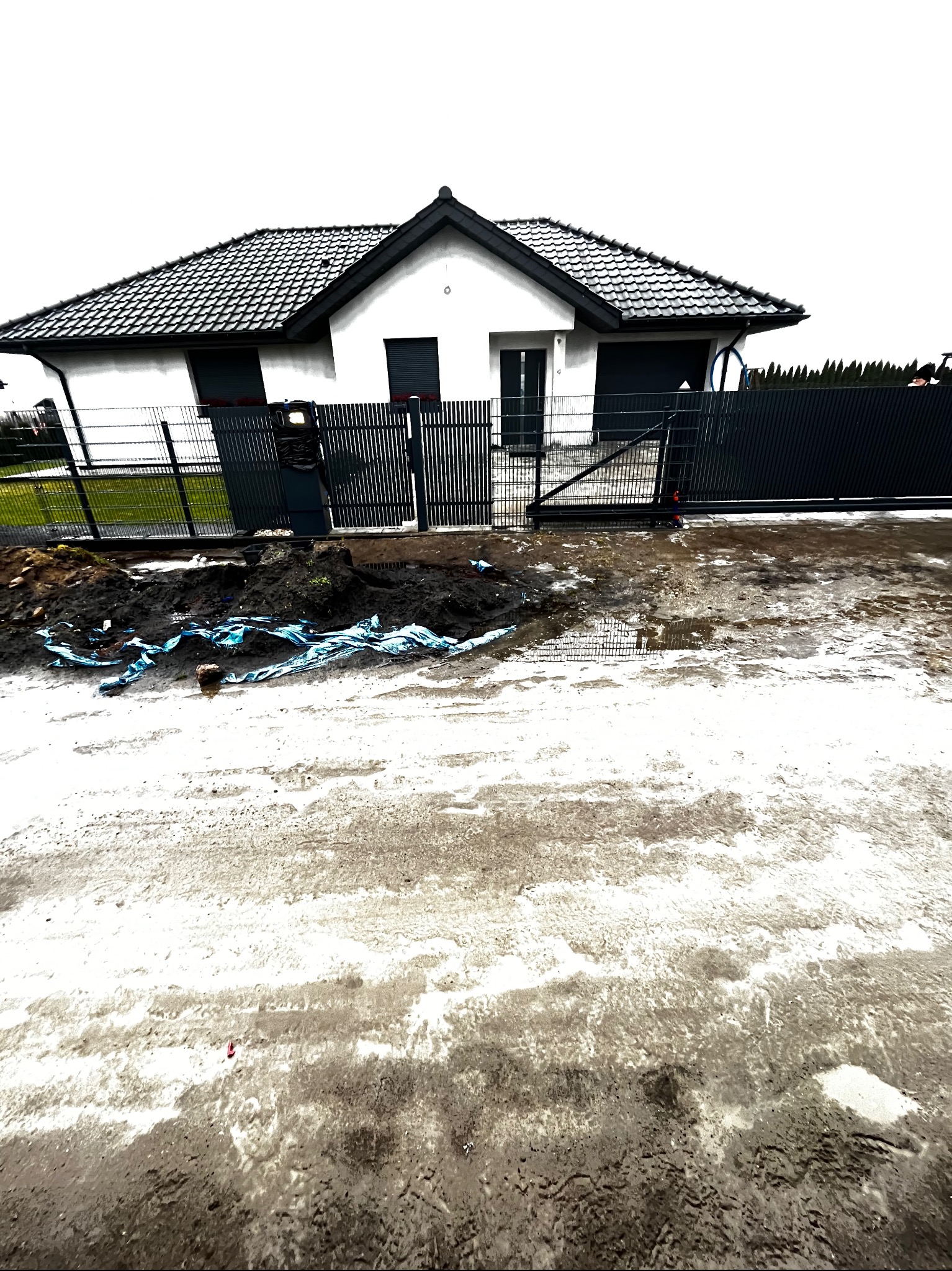 Nowoczesny dom jednorodzinny z grafitowym ogrodzeniem panelowym i bramą wjazdową. Widoczne prace ziemne przed posesją. Ujęcie z dołu, podkreślające bryłę budynku.