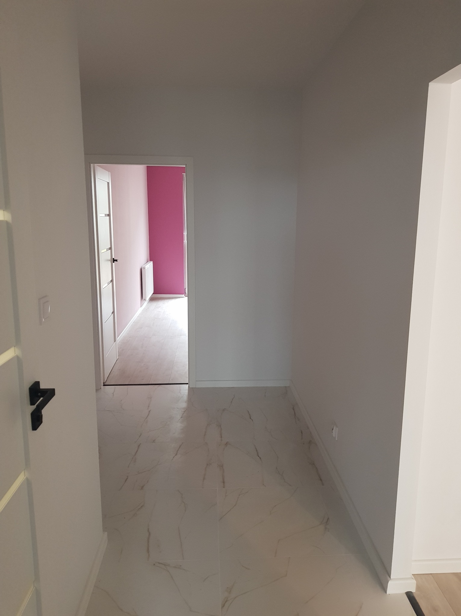Korytarz z białymi ścianami i marmurową podłogą, widok na pokój z różową ścianą i białym grzejnikiem.