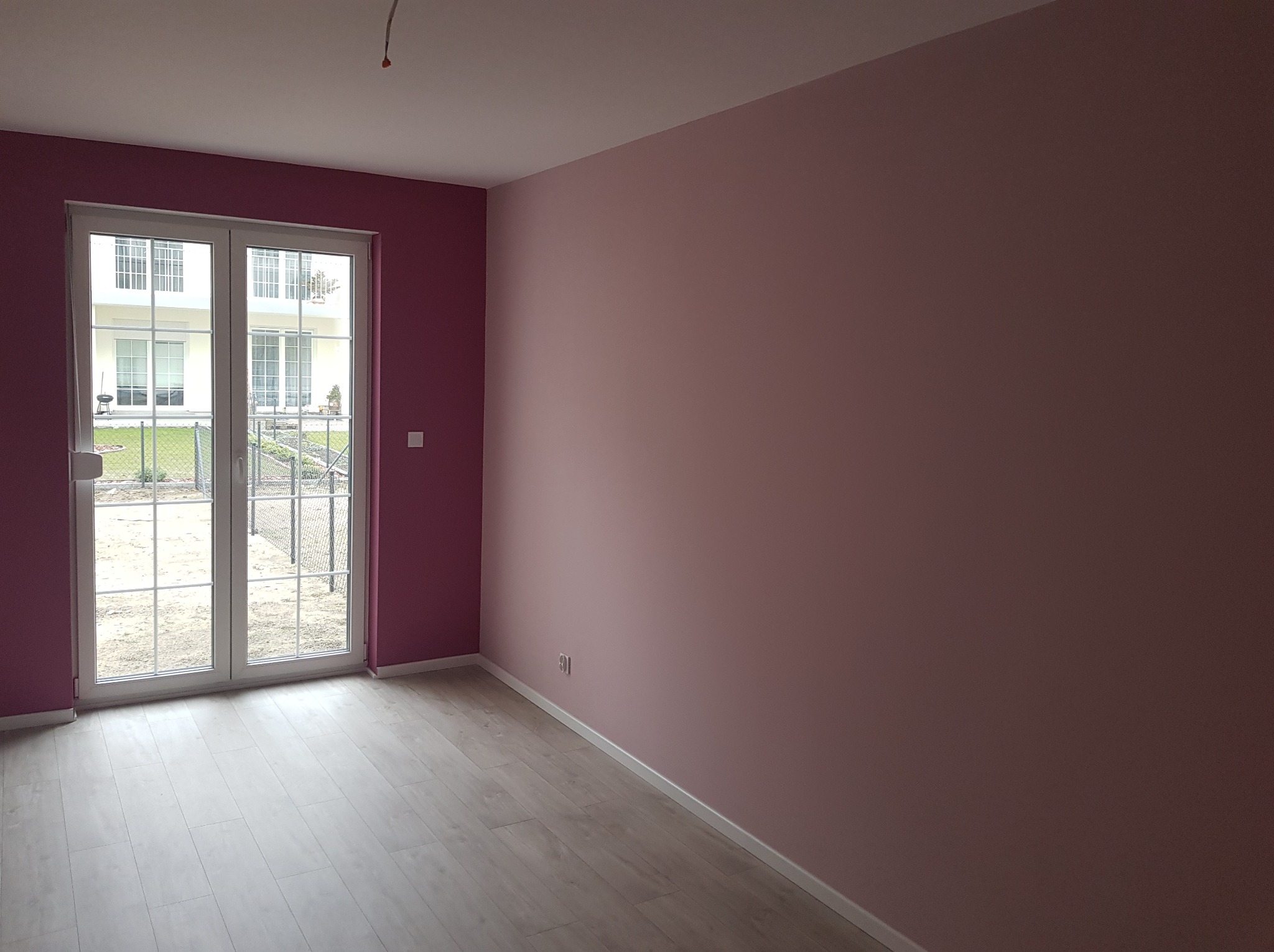 Pusty pokój po malowaniu ścian na odcienie różu, widoczne białe listwy przypodłogowe i panele podłogowe, z wyjściem na balkon z widokiem na ogród.
