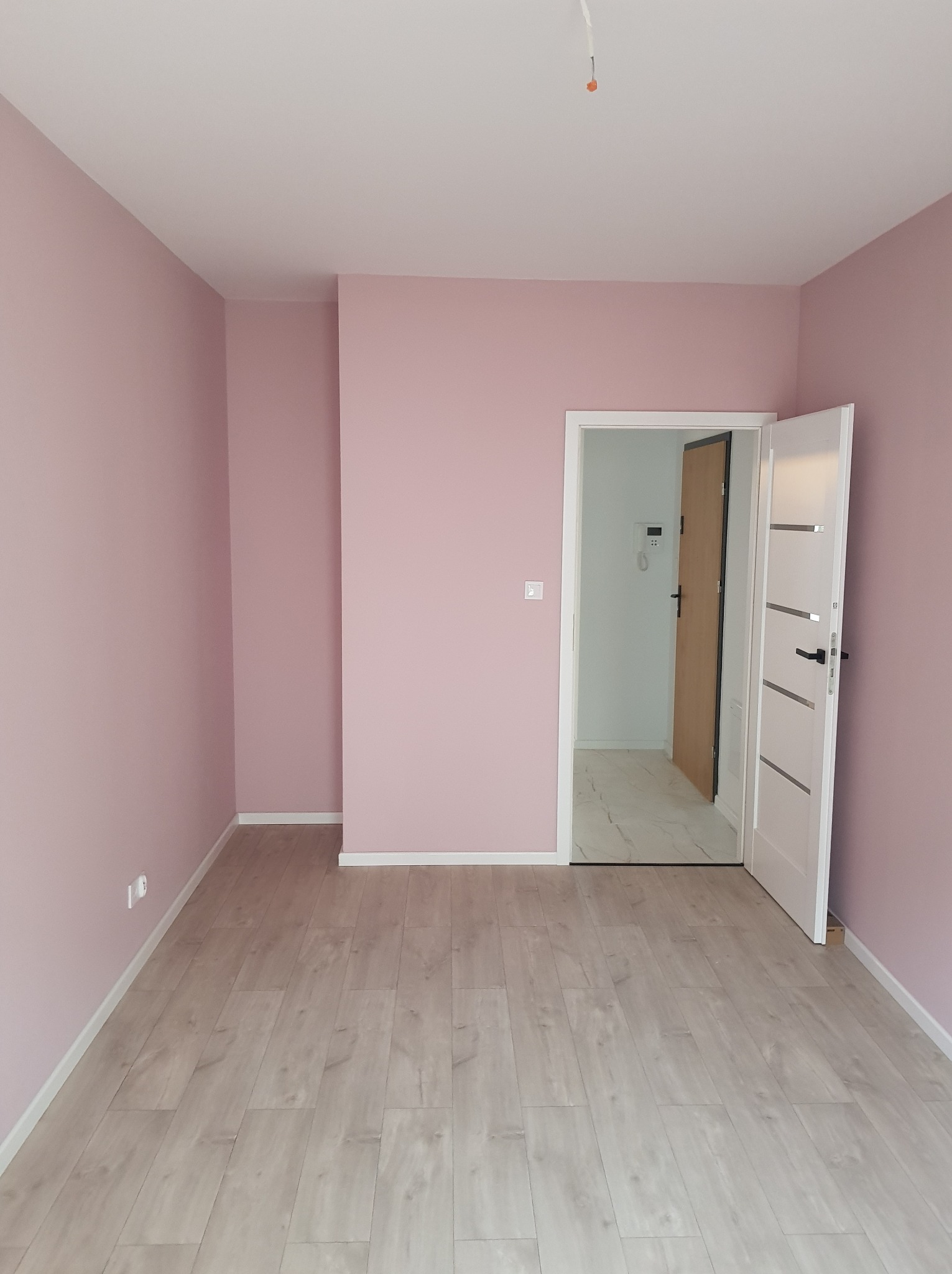 Pusty pokój z pomalowanymi na różowo ścianami, jasną podłogą i otwartymi białymi drzwiami z poziomymi srebrnymi wstawkami.