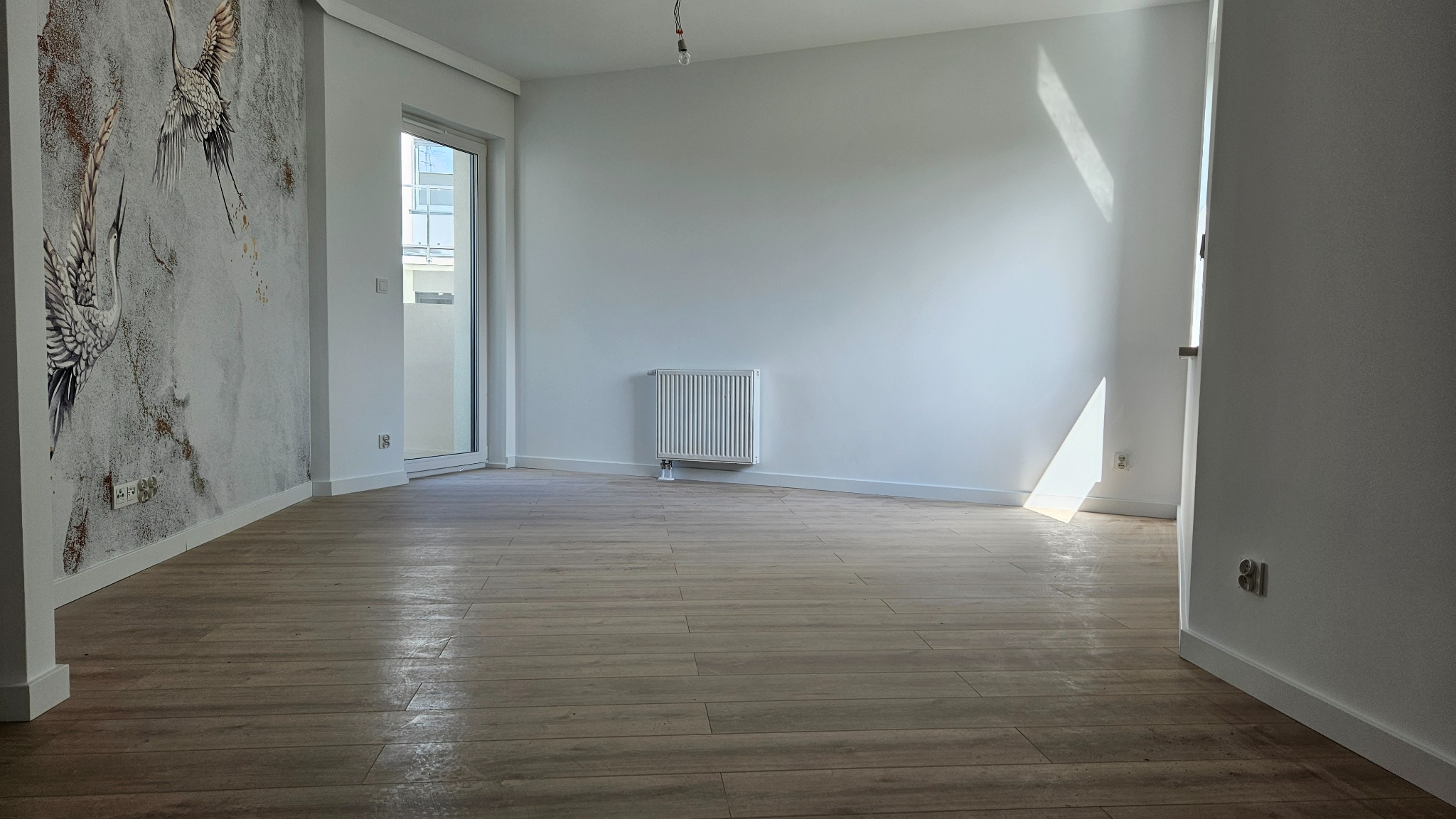 Jasne, puste pomieszczenie z drewnianą podłogą, świeżo pomalowanymi ścianami, na jednej ze ścian widoczna tapeta z motywem żurawi, grzejnik przy ścianie, doświetlone promieniami słonecznymi...