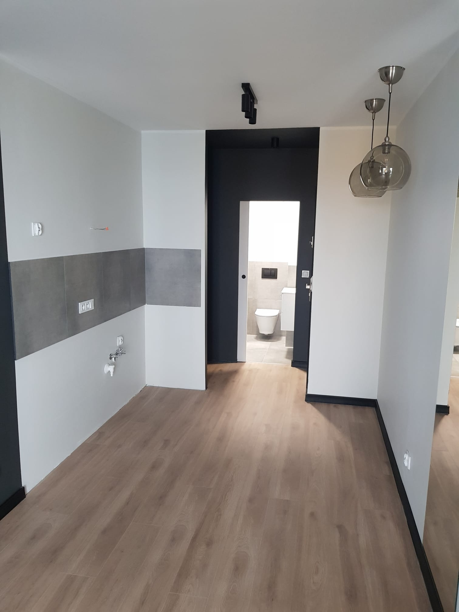 Wnętrze mieszkania w trakcie wykończenia: pomalowane ściany na biało, częściowo położone szare płytki w kuchni, widoczne wyprowadzenia instalacji, drewnopodobna podłoga i otwarte drzwi do łazienki...