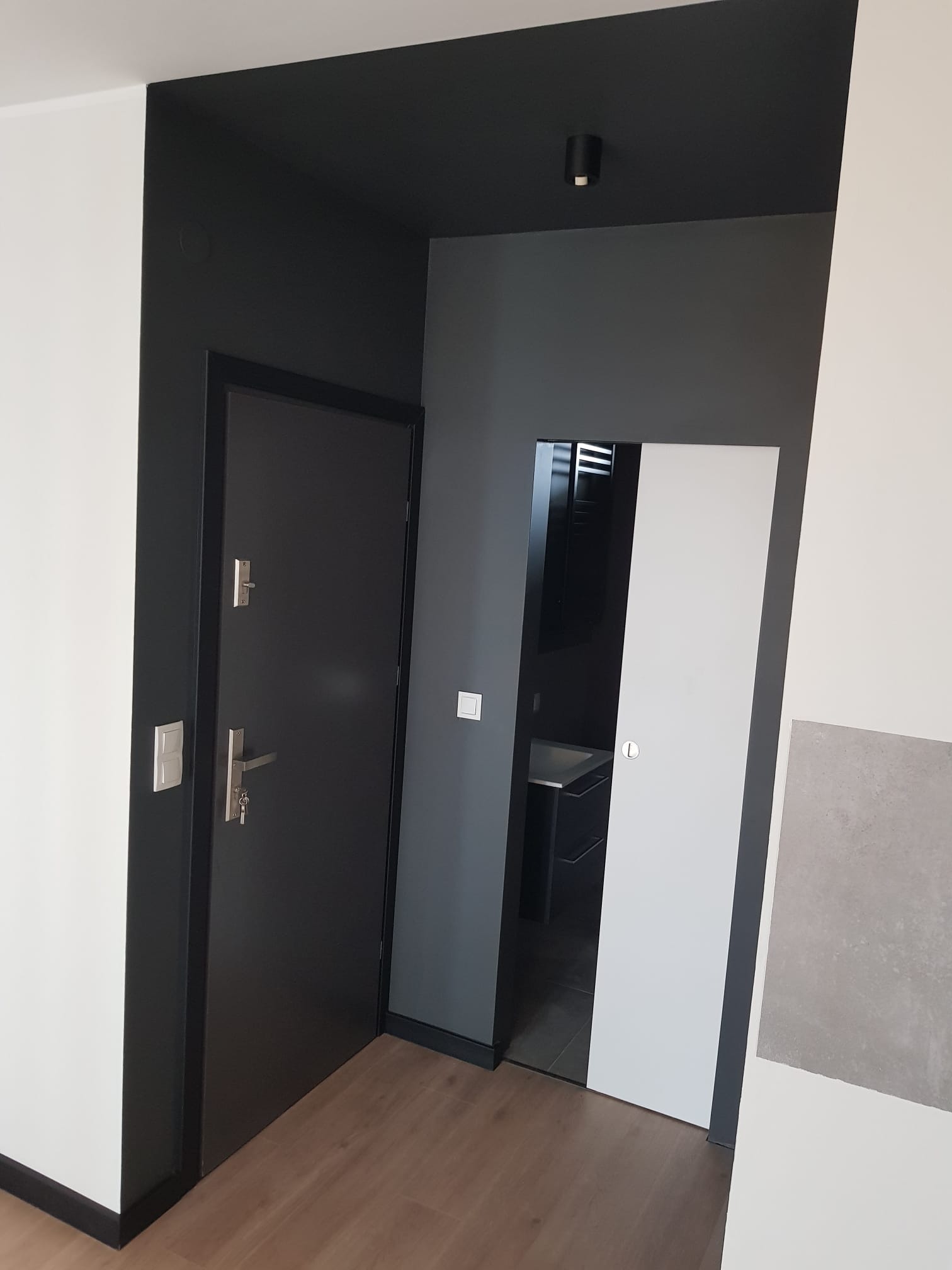 Wnętrze po remoncie: kontrastowe zestawienie białych ścian z czarną ramą drzwi wejściowych i szarym tłem, widoczna przesuwana biała ściana i drewniana podłoga.