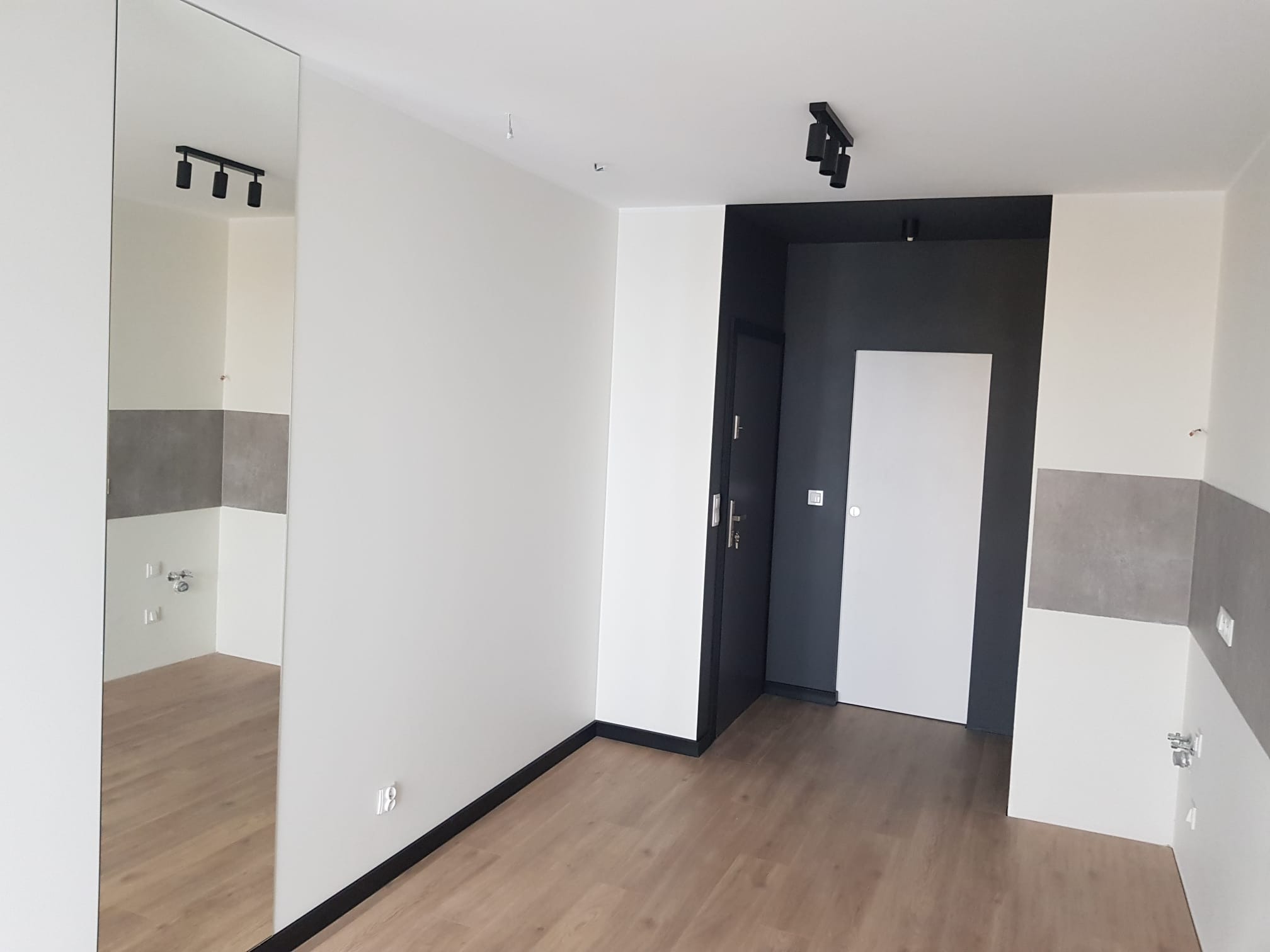 Wnętrze po malowaniu: białe ściany z szarym pasem dekoracyjnym, drewniana podłoga, czarne akcenty w postaci oświetlenia i futryn drzwiowych, lustro odbijające fragment pomieszczenia.