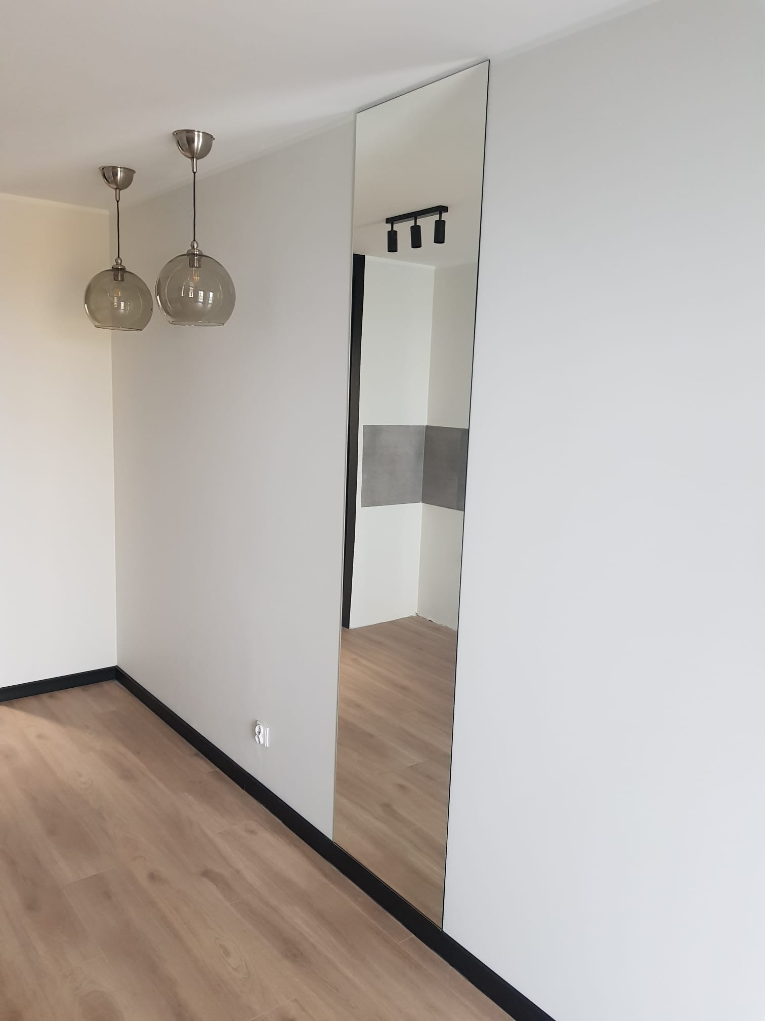 Pomalowane na biało wnętrze z drewnopodobną podłogą, wysokim lustrem na ścianie, odbijającym fragment kuchni, oraz dwoma szklanymi lampami wiszącymi.