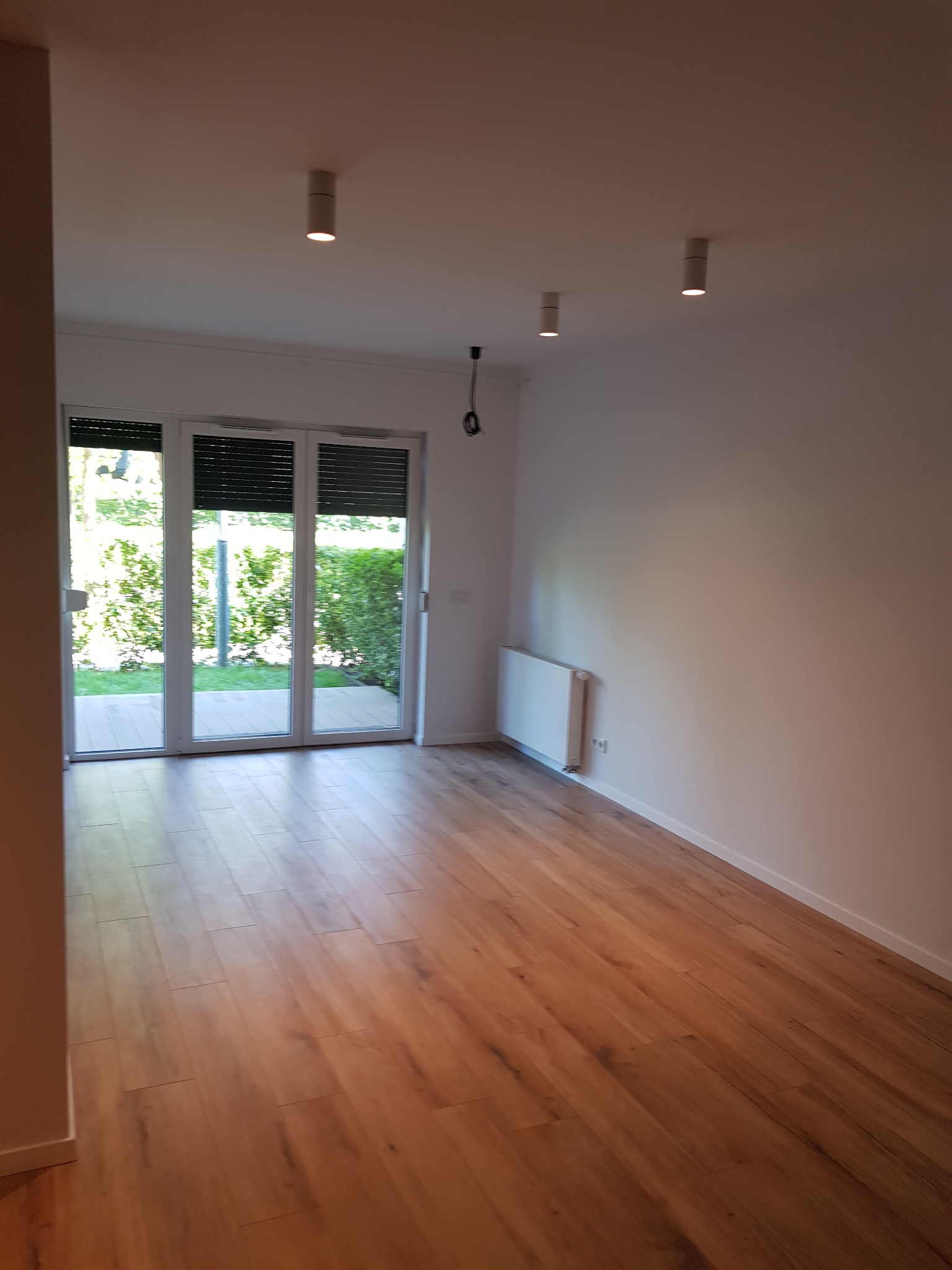 Pusty, jasny pokój z drewnianą podłogą, białymi ścianami i trzema punktowymi lampami sufitowymi; widok na ogród przez przeszklone drzwi balkonowe z żaluzjami.