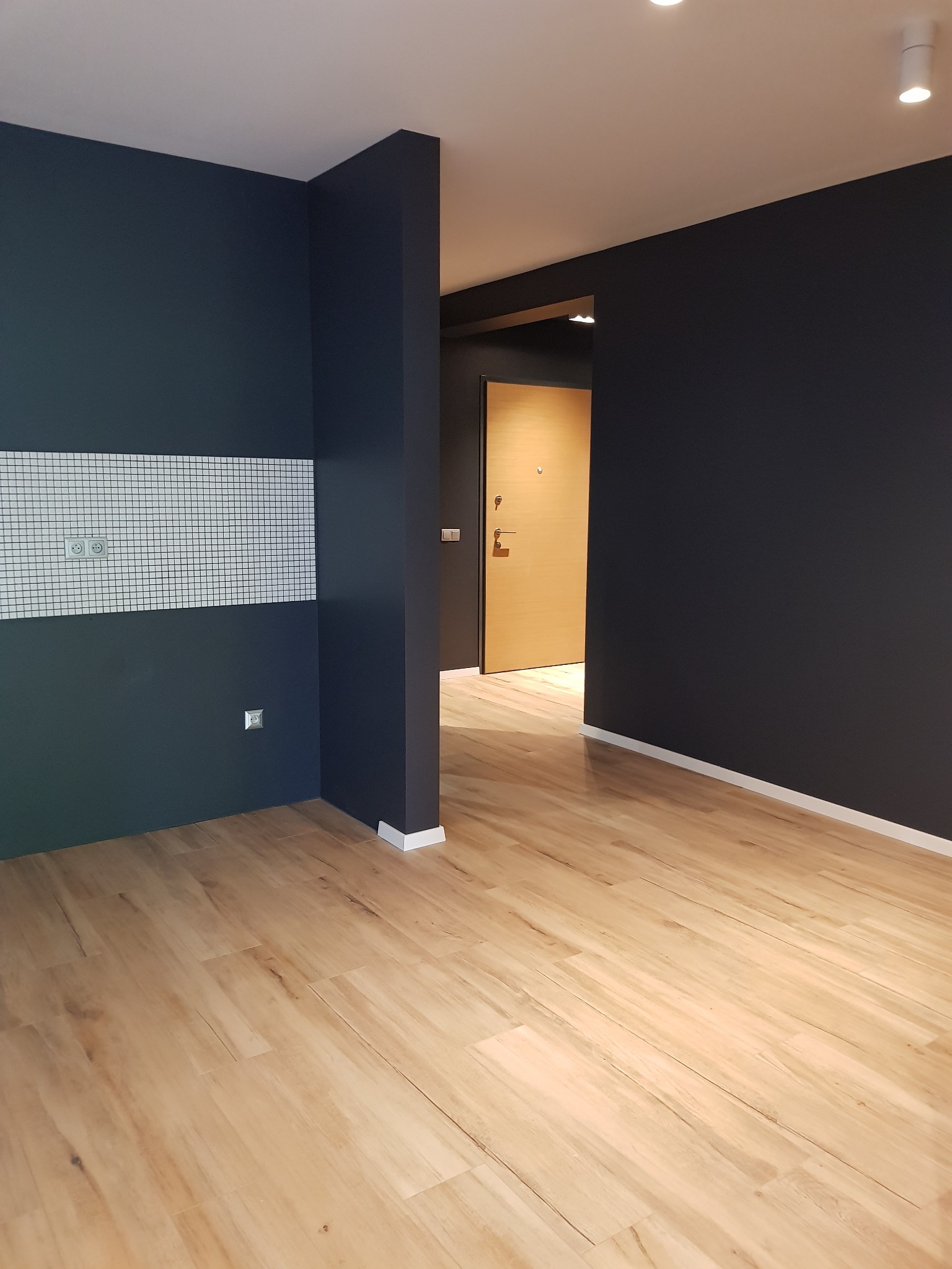Świeżo pomalowane na ciemny granatowo-szary kolor ściany w pustym pokoju z drewnianą podłogą i białymi listwami przypodłogowymi, widoczny fragment ściany z białymi płytkami mozaikowymi i gniazdkami.