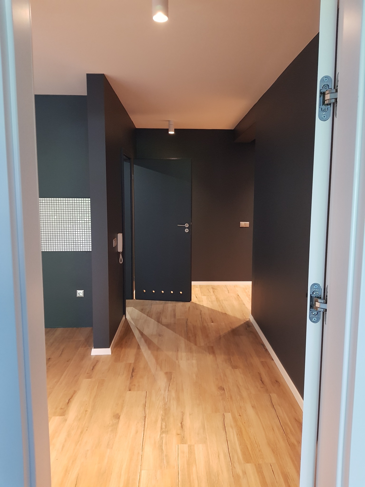 Wnętrze po remoncie: ciemnoszare ściany, jasna drewniana podłoga, widok zza białych drzwi.