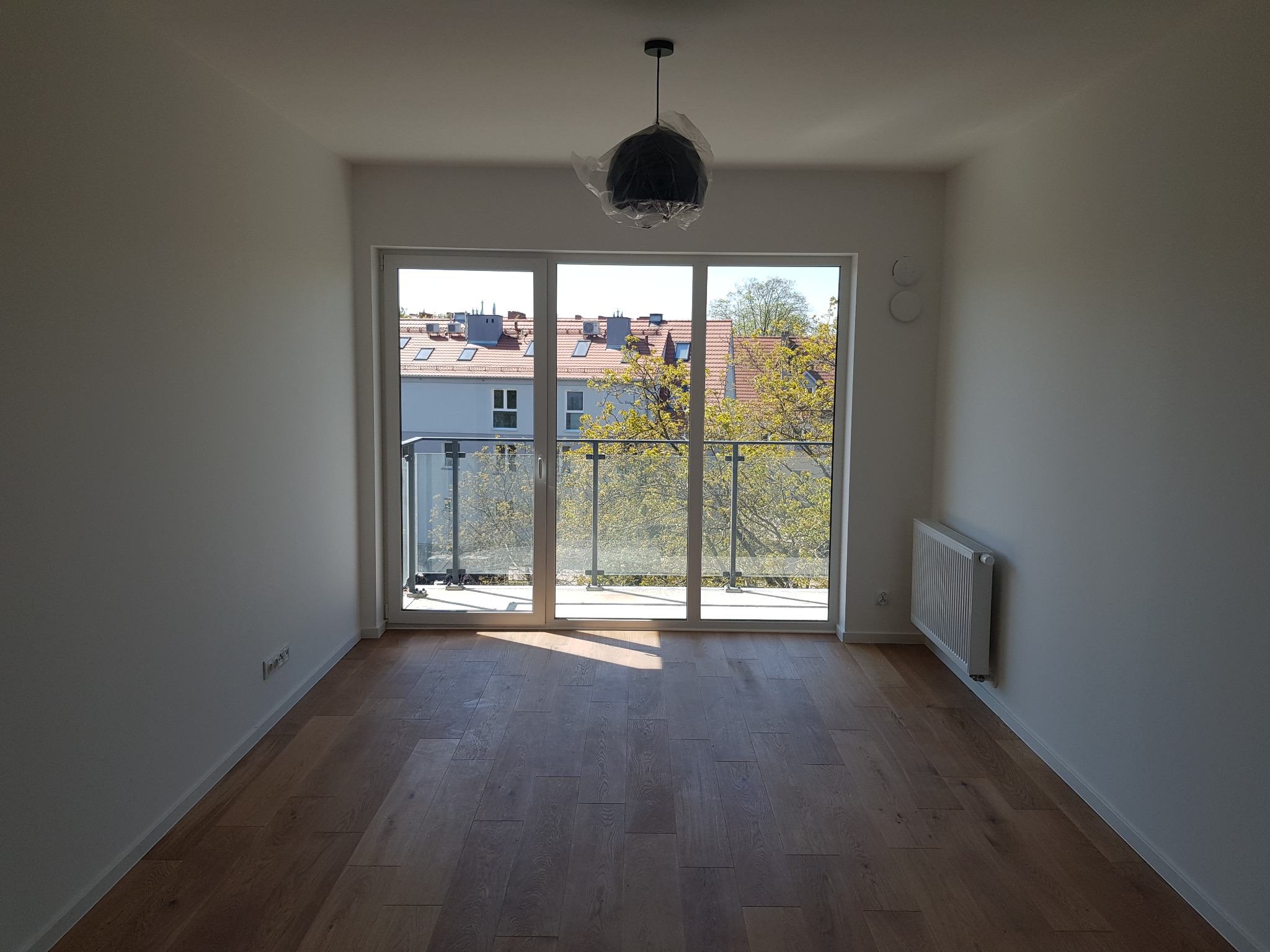 Puste, jasne pomieszczenie z parkietem, balkonem i grzejnikiem; lampa wisząca zabezpieczona folią ochronną.