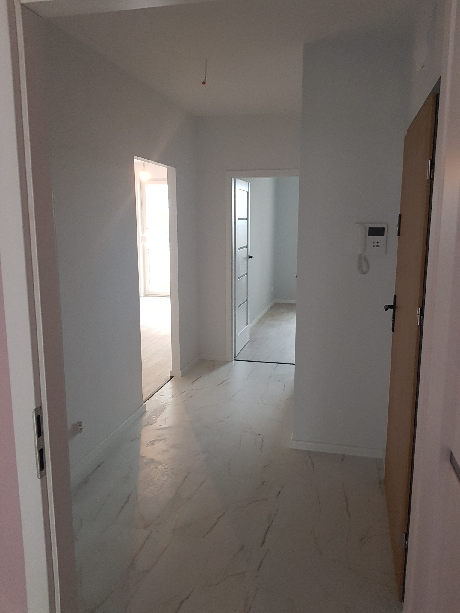 Wnętrze korytarza po remoncie, ściany pomalowane na biało, podłoga wyłożona jasnymi płytkami imitującymi marmur, widoczne drzwi do innych pomieszczeń.