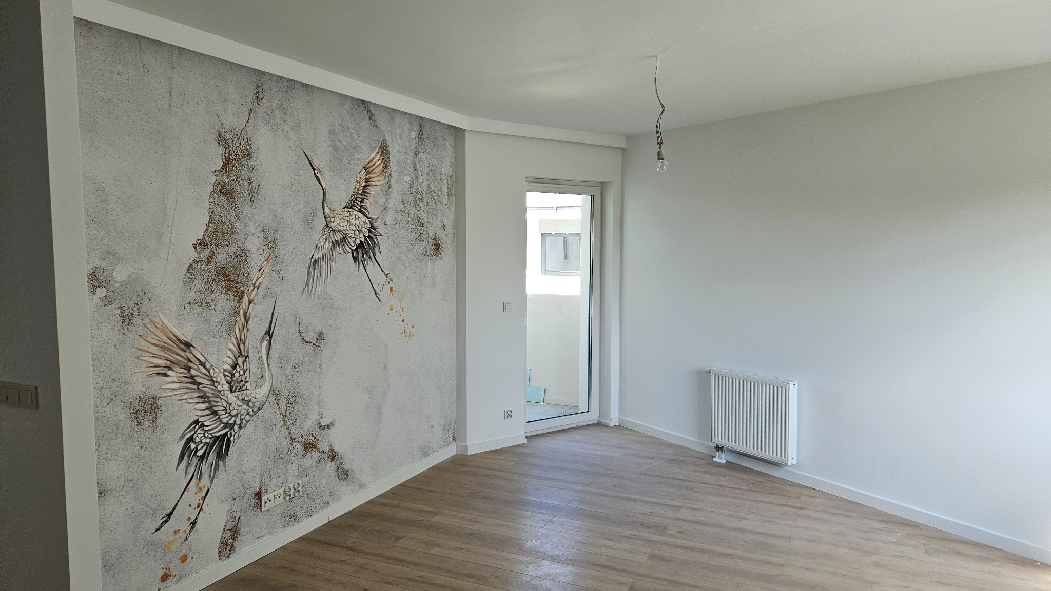 Wnętrze pokoju po remoncie: ściana z szarą tapetą w motywie żurawi, drewniana podłoga, biały grzejnik i surowa żarówka zwisająca z sufitu.