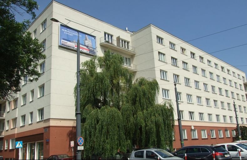 Wielopiętrowy budynek mieszkalny z odnowioną elewacją w kolorze kremowym, częściowo zasłonięty przez rozłożyste drzewo, z widocznym fragmentem reklamy na ścianie i zaparkowanymi samochodami.