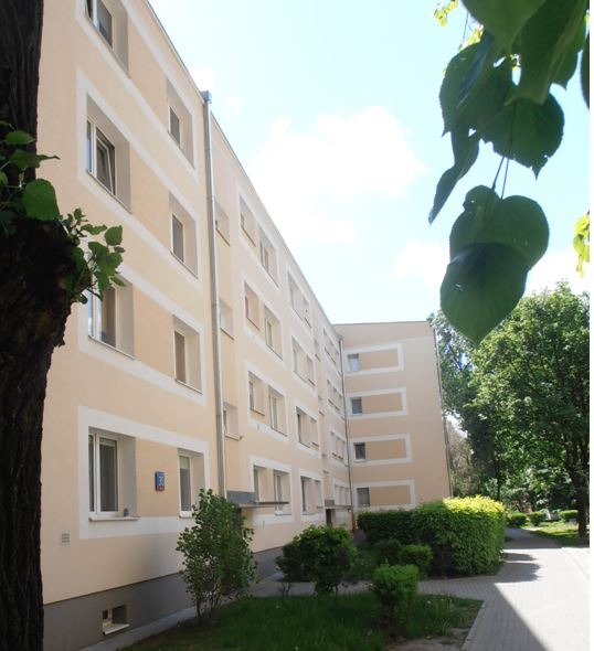 Wielorodzinny budynek po renowacji elewacji w odcieniach beżu i kremu, z widocznymi obramowaniami okien. Na pierwszym planie zieleń i fragment drzewa.