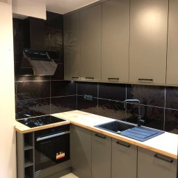 Meble Rzeszow - Nowoczesna kuchnia z szarymi szafkami, drewnianym blatem, czarnym zlewem i płytą indukcyjną, czarne błyszczące płytki na ścianie, oświetlenie punktowe.