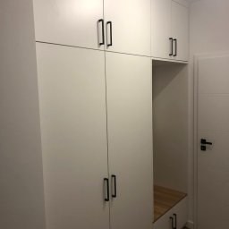 Meble Rzeszow - Biała szafa wnękowa z czarnymi, minimalistycznymi uchwytami, zawierająca otwartą wnękę z drewnianym siedziskiem, przylegająca do białych drzwi z poziomymi przeszyciami.