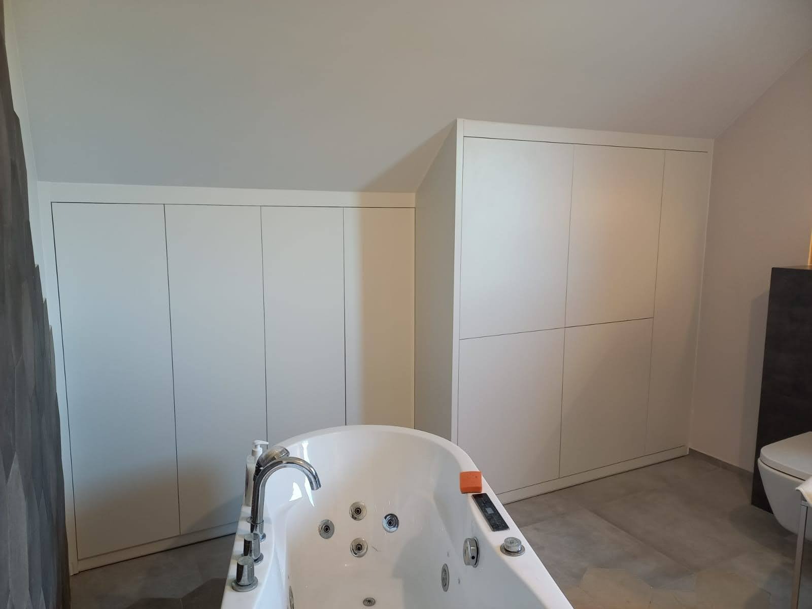 Białe szafy wnękowe na poddaszu w łazience z wanną jacuzzi, widoczne minimalistyczne fronty i skośny sufit.