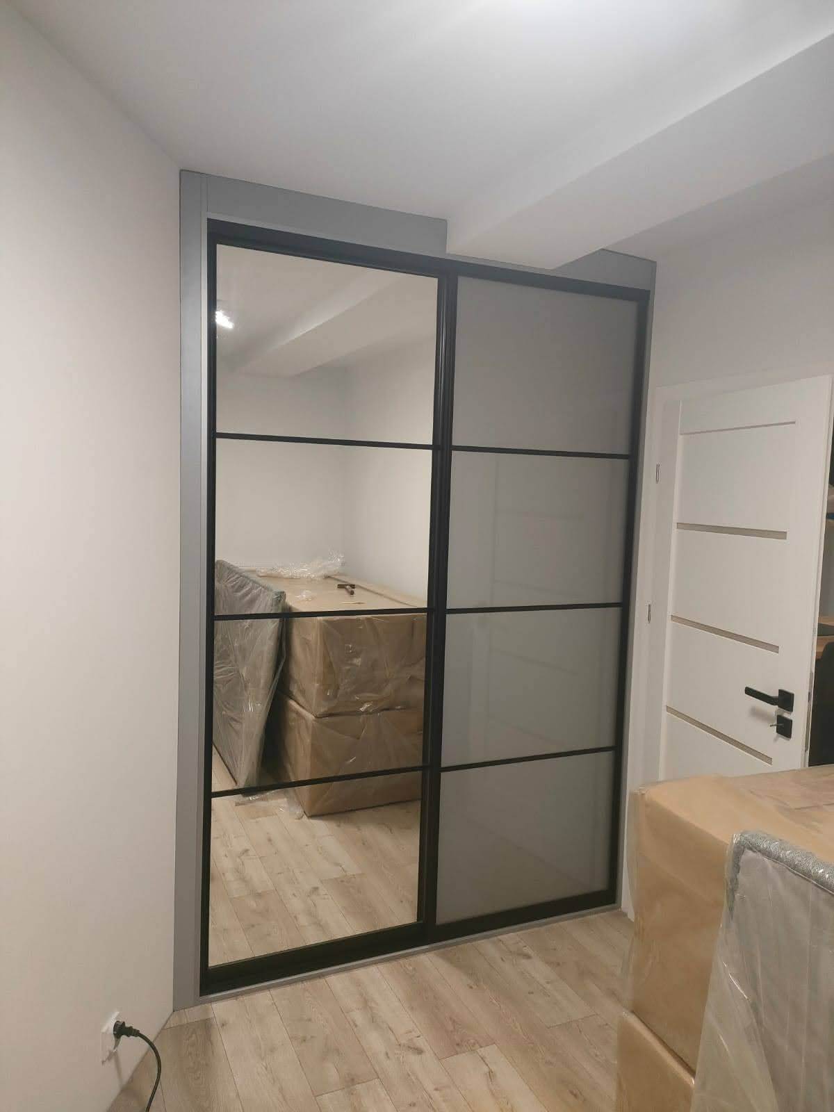 Nowoczesna szafa wnękowa z przesuwnymi drzwiami, czarne ramy, częściowo lustrzane, widoczne kartony wewnątrz, jasne panele podłogowe.