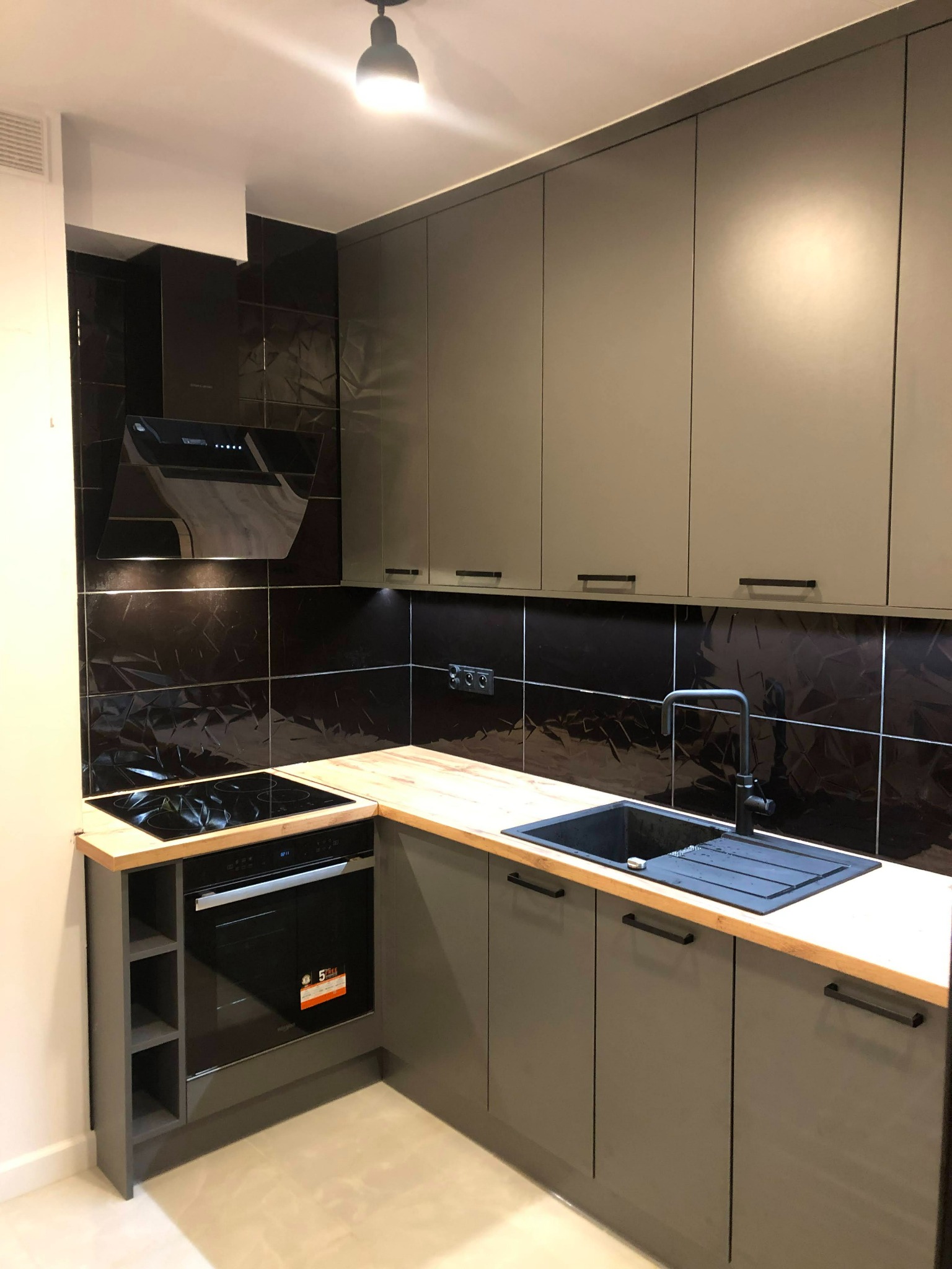 Nowoczesna kuchnia z szarymi szafkami, drewnianym blatem, czarnym zlewem i płytą indukcyjną, czarne błyszczące płytki na ścianie, oświetlenie punktowe.