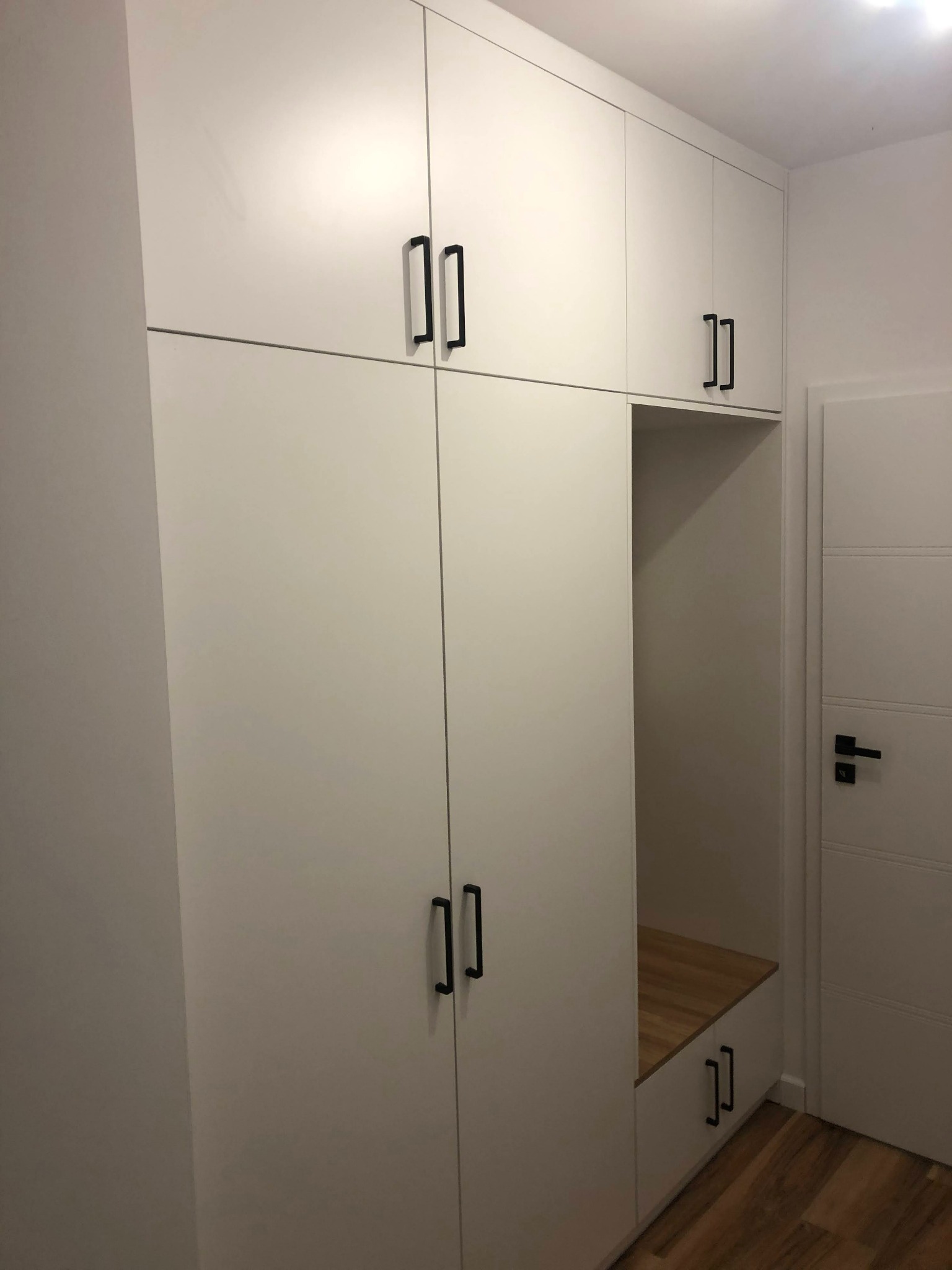 Biała szafa wnękowa z czarnymi, minimalistycznymi uchwytami, zawierająca otwartą wnękę z drewnianym siedziskiem, przylegająca do białych drzwi z poziomymi przeszyciami.