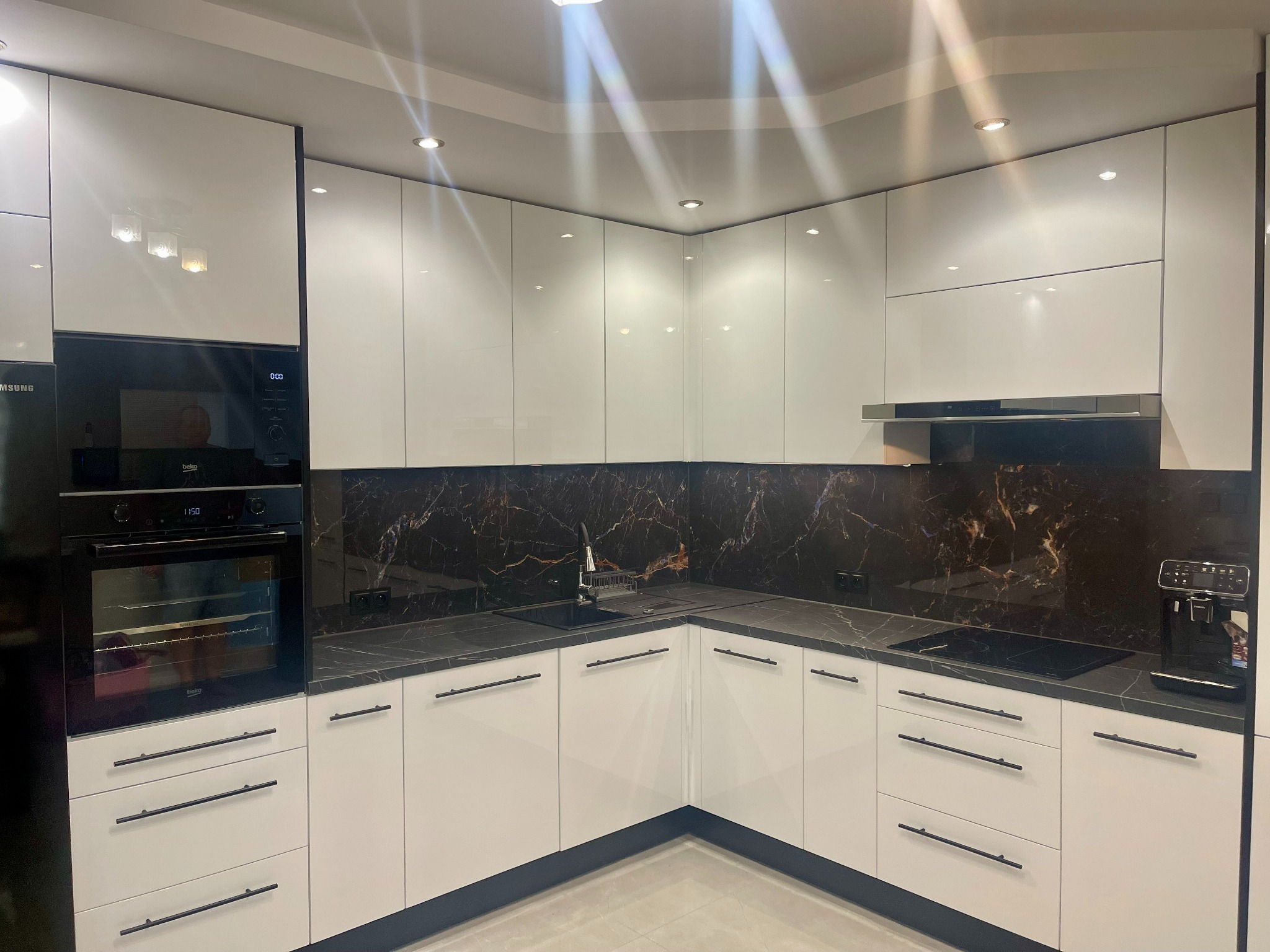 Nowoczesna kuchnia w kształcie litery L z białymi, lakierowanymi frontami, czarnym blatem imitującym marmur oraz zabudowanym piekarnikiem i ekspresem do kawy.