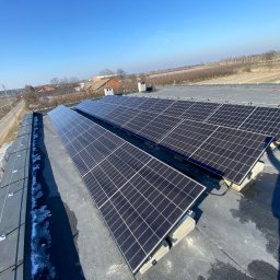 Bonner Firma Instalacyjna - Panele fotowoltaiczne zamontowane na płaskim dachu budynku gospodarczego, widoczne palety z materiałami budowlanymi i resztki śniegu wzdłuż krawędzi dachu, w tle wiejski krajobraz i błękitne niebo.