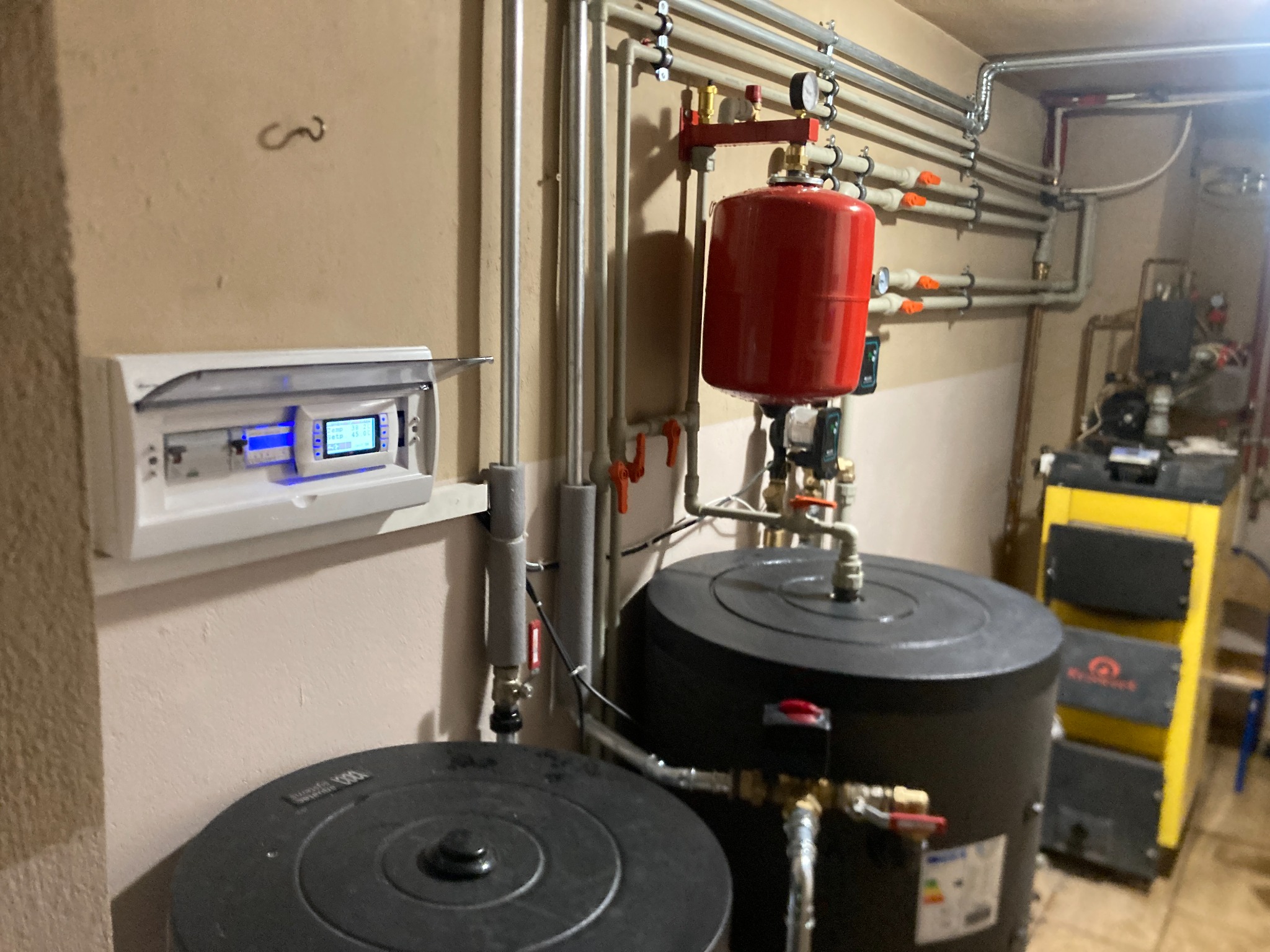 Kotłownia z dwoma czarnymi zbiornikami, czerwonym zasobnikiem, rurami instalacji grzewczej i żółto-czarnym piecem w tle, widoczny panel sterowania na ścianie.