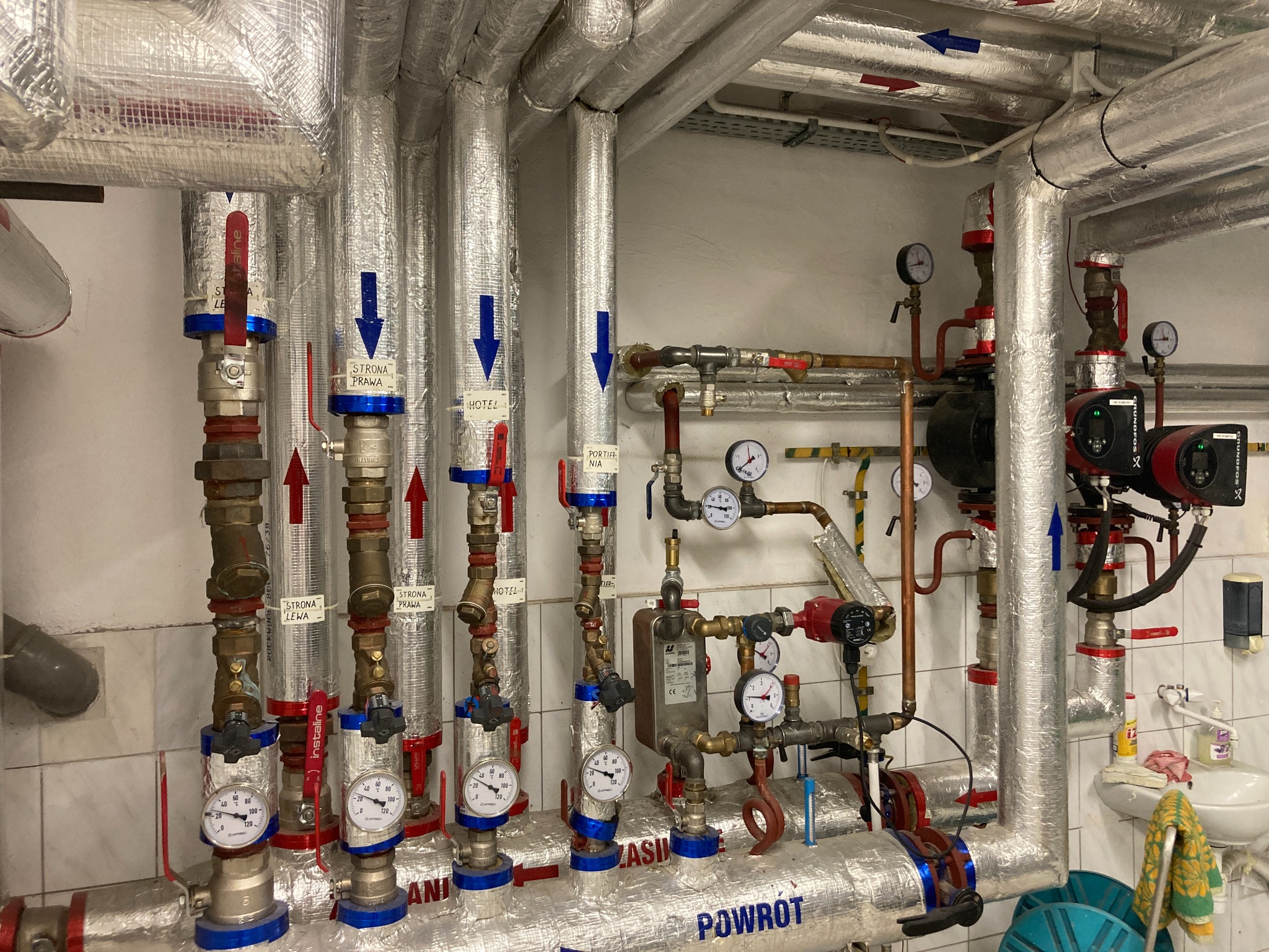 Szczegółowe ujęcie węzła cieplnego z rurami w izolacji aluminiowej, zaworami oznaczonymi kierunkiem przepływu i manometrami, widoczne elementy instalacji centralnego ogrzewania w pomieszczeniu...