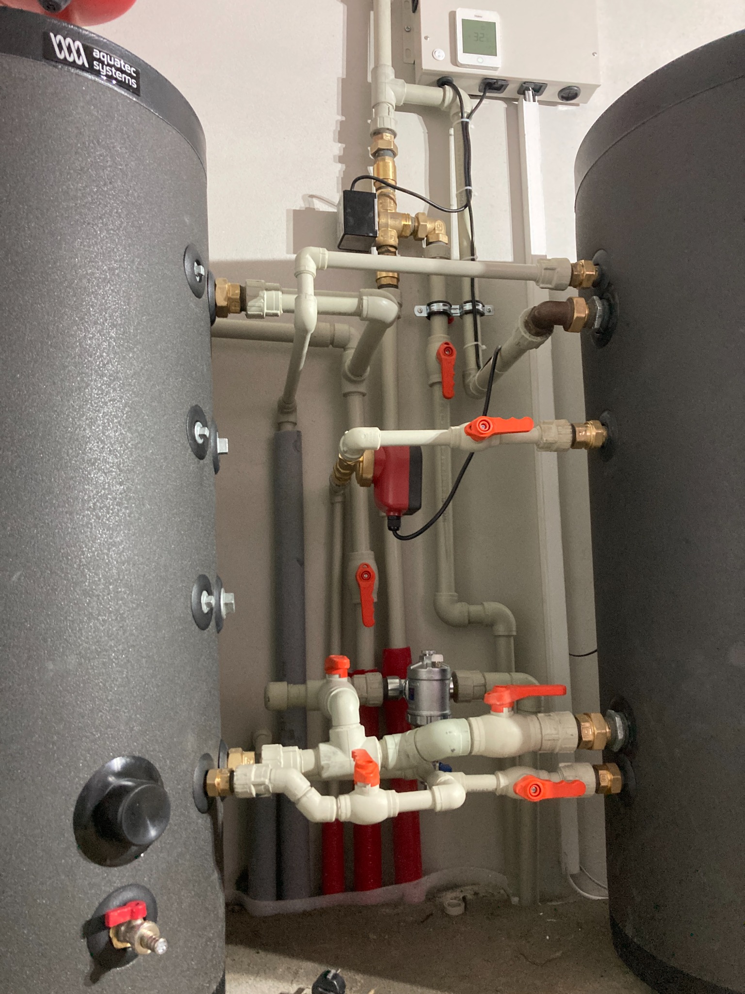 Dwa szare zbiorniki Aquatec Systems połączone rurami z czerwonymi zaworami, widoczny sterownik temperatury na ścianie.