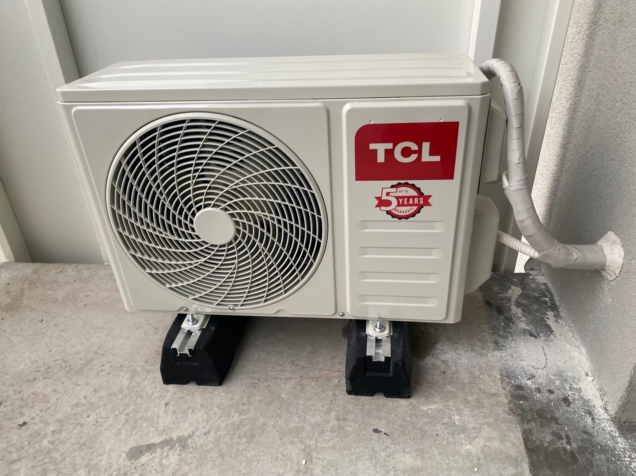 Biała jednostka zewnętrzna klimatyzacji TCL z widocznym logo i naklejką 5 lat gwarancji, zamontowana na czarnych podstawkach antywibracyjnych z aluminiowymi wspornikami na szarym betonowym podłożu,...