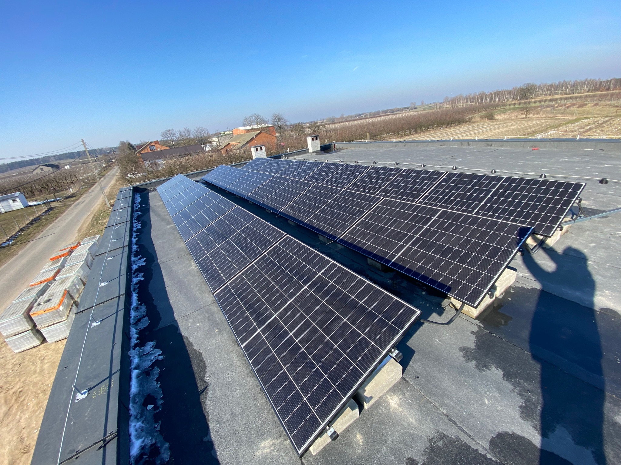 Panele fotowoltaiczne zamontowane na płaskim dachu budynku gospodarczego, widoczne palety z materiałami budowlanymi i resztki śniegu wzdłuż krawędzi dachu, w tle wiejski krajobraz i błękitne niebo.