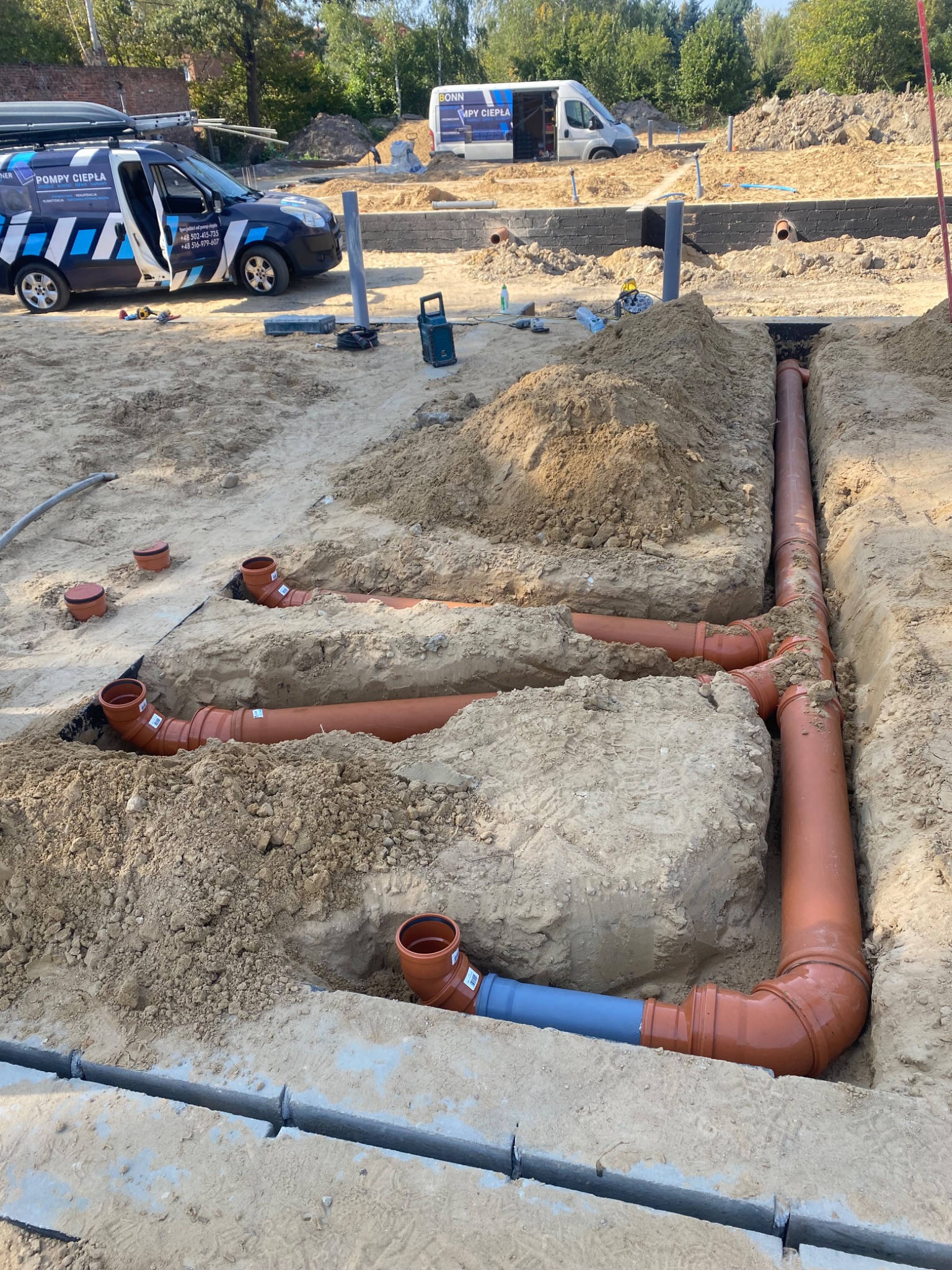 Wykonanie instalacji z pomarańczowych rur w wykopie ziemnym, widoczny fragment niebieskiej rury, na drugim planie samochody dostawcze firmy od OZE