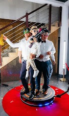 Trzy osoby, w tym dziecko, pozują na obrotowej platformie z rekwizytami na tle schodów z metalową balustradą, prawdopodobnie podczas imprezy.