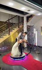 Para w strojach ślubnych tańczy na platformie obrotowej, otoczona konfetti, na czerwonym dywanie, z widocznym oświetleniem pierścieniowym i złotym tłem.