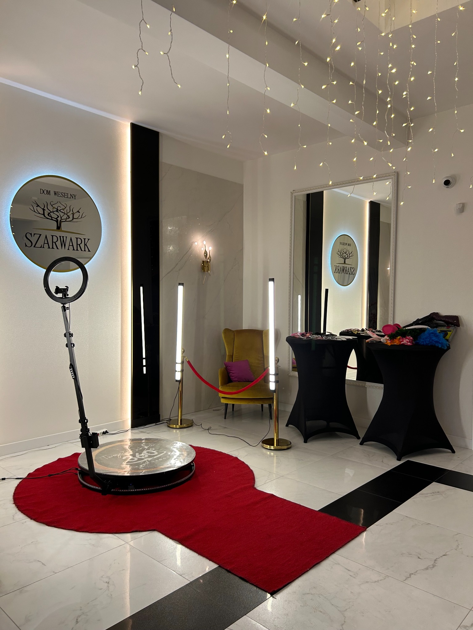 Wnętrze z okrągłą platformą do nagrywania filmów 360 stopni, otoczoną czerwonym dywanem, oświetloną lampami LED, z rekwizytami na stolikach w tle i dekoracyjnymi lampkami na suficie.