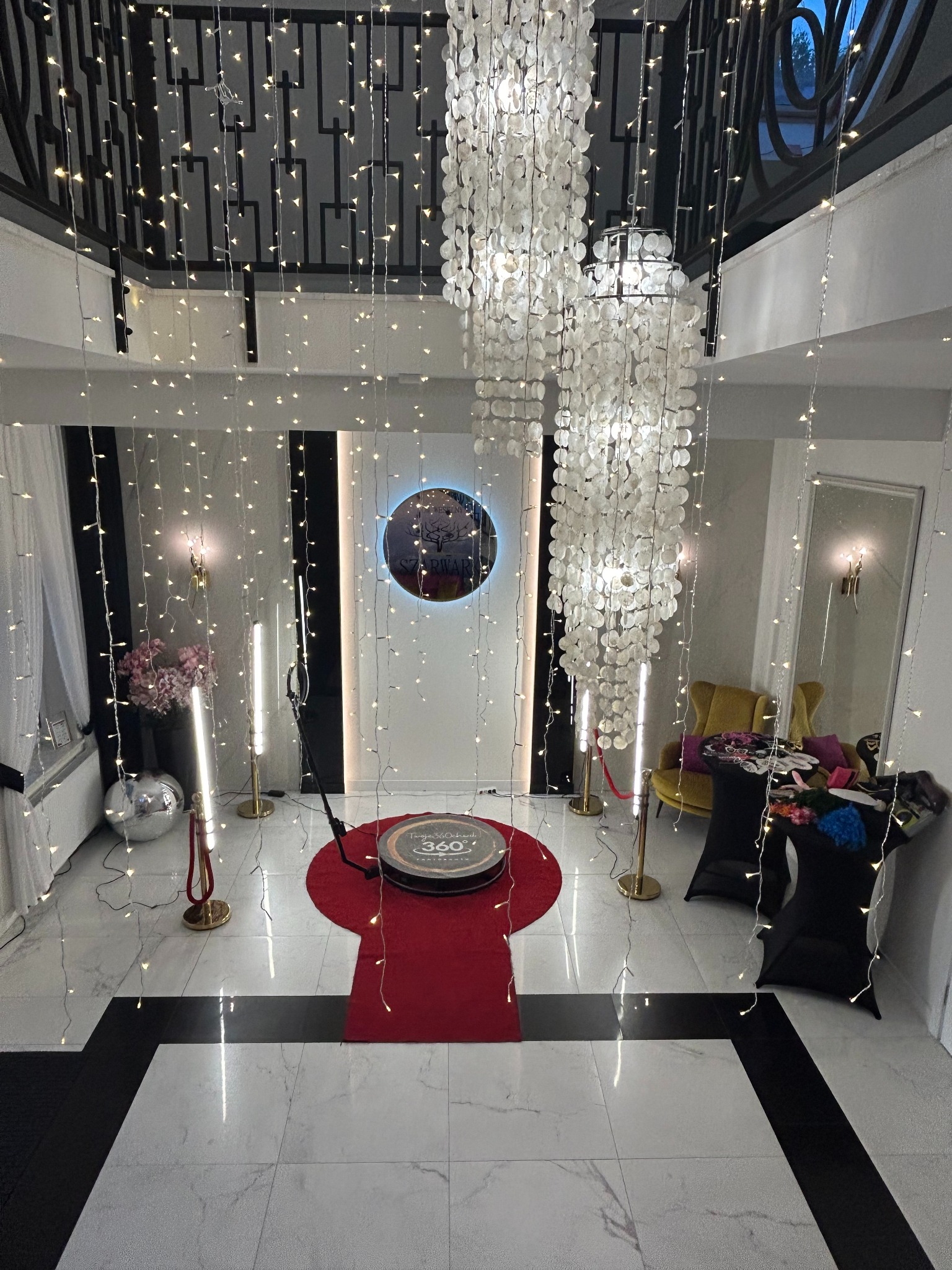 Wnętrze z okrągłą platformą na czerwonym dywanie, otoczoną lampkami, dekoracjami i rekwizytami, tworzące elegancką przestrzeń do zdjęć.