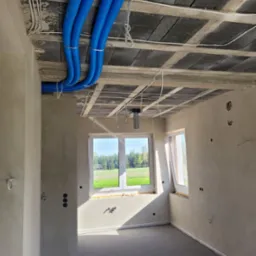 Wnętrze w stanie surowym: widoczne niebieskie rury instalacyjne pod sufitem, wystające przewody elektryczne, okna z widokiem na zieleń. Stan deweloperski.