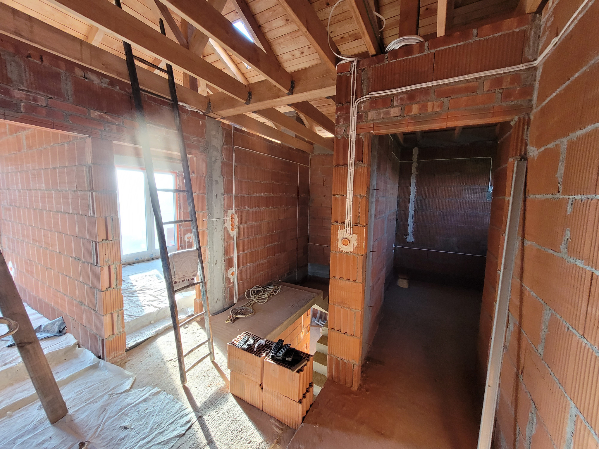 Wnętrze w trakcie budowy z widoczną instalacją elektryczną: kable prowadzone po ścianach z czerwonej cegły, puszki pod gniazdka i drewniana konstrukcja dachu.
