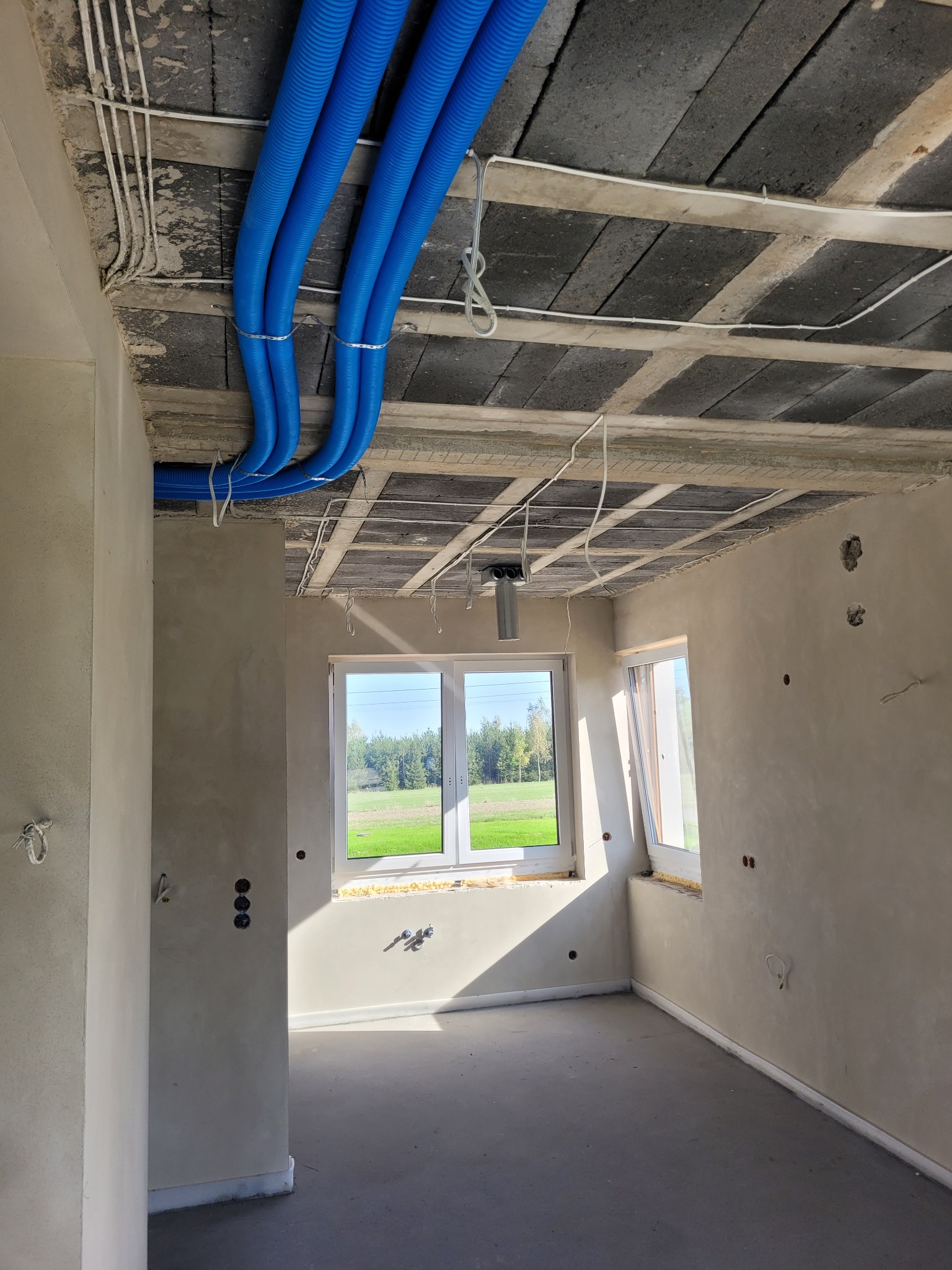 Wnętrze w stanie surowym: widoczne niebieskie rury instalacyjne pod sufitem, wystające przewody elektryczne, okna z widokiem na zieleń. Stan deweloperski.
