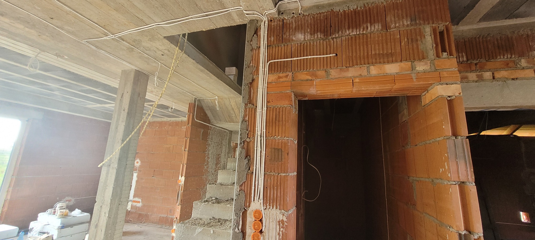 Instalacja elektryczna w budynku w stanie surowym: białe kable prowadzone po suficie i ścianach z czerwonej cegły, widoczne puszki elektryczne. Budowa domu w trakcie prac.